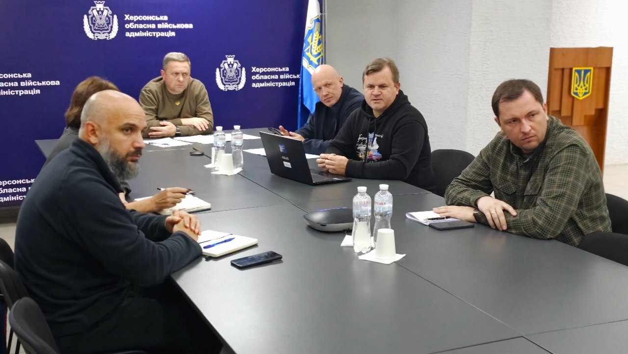 На офіційній зустрічі за великим сірим столом сидить група посадовців, які обговорюють програми підтримки ветеранів та підготовку до опалювального сезону. По центру на задньому плані прямо під синім банером ХОВА сидить перший заступник начальника Херсонської ОВА Дмитро Бутрій у кофті кольору хакі. Ліворуч від Бутрія сидить заступниця Херсонського міського голови з питань діяльності виконавчих органів ради Наталія Чорненька, яка частково потрапила в лівий край кадру. Праворуч від Бутрія сидить заступник начальника ХОВА Євгеній Ігнатенко у темно-синій кофті. Праворуч від Ігнатенка перебуває заступник начальника ХОВА Володимир Клюцевський у чорному худі з малюнком, перед яким стоїть відкритий чорний ноутбук. Праворуч від Клюцевського сидить директор Департаменту з питань цивільного захисту та оборонної роботи Херсонської ОДА Євген Пісоцький у картатій сорочці кольору хакі. На передньому плані ліворуч у профіль сидить начальник управління з питань децентралізації та розвитку громад Херсонської ОДА Юліан Лучко — чоловік із бородою у чорній кофті з відкритим блокнотом. На столі перед учасниками розставлені пляшки з водою, паперові склянки та лежить канцелярське приладдя. Ззаду на стіні зали розташовані сині банери з гербами та написами «Херсонська обласна військова адміністрація», а у верхньому правому кутку помітно дерев'яну трибуну із золотим тризубом.