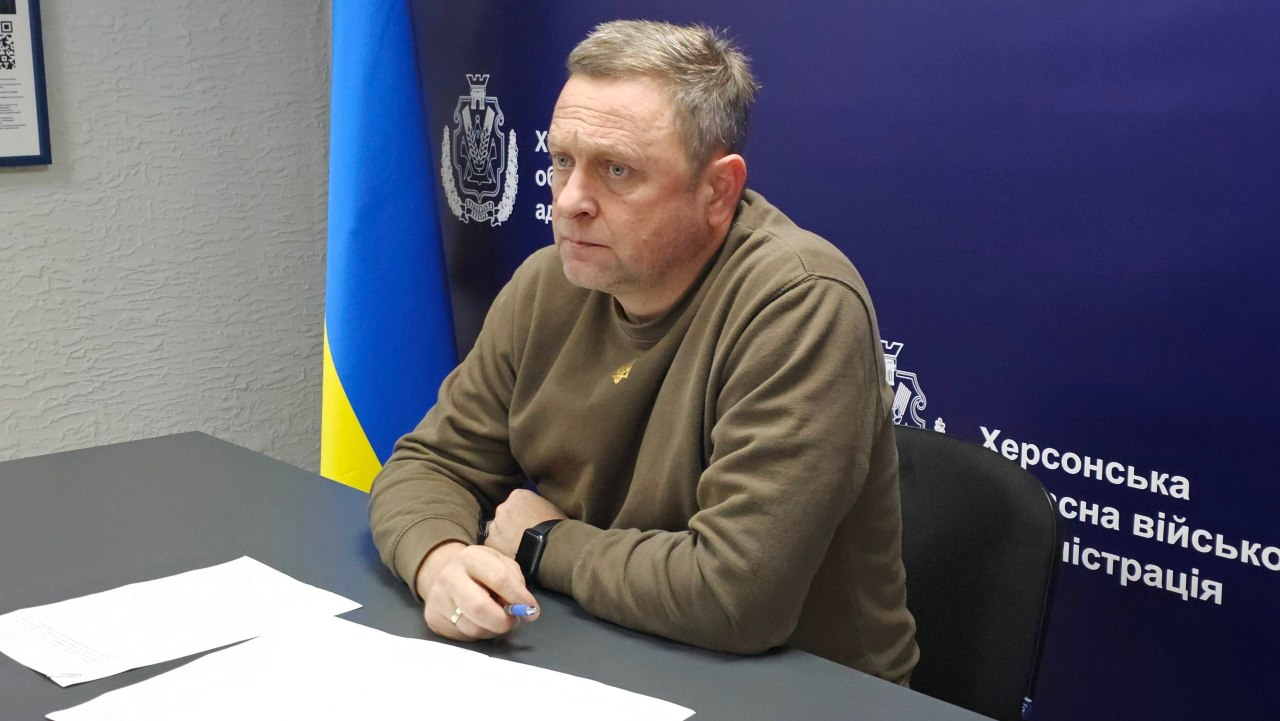 У кабінеті за сірим робочим столом великим планом зафіксовано першого заступника начальника Херсонської ОВА Дмитра Бутрія, який головує на нараді щодо оновлення місцевих програм підтримки ветеранів та підготовки до опалювального сезону. Він одягнений у кофту (світшот) кольору хакі з невеликим патріотичним принтом на грудях, носить смартгодинник на лівій руці та тримає синю кулькову ручку правою рукою, уважно дивлячись у бік співрозмовників. Перед ним на столі розкладено білі робочі аркуші паперу з документами. На задньому плані ліворуч стоїть державний прапор України, а праворуч розташований великий темно-синій банер із офіційним гербом та білими написами, серед яких чітко видно частину тексту «Херсонська обласна військова адміністрація».