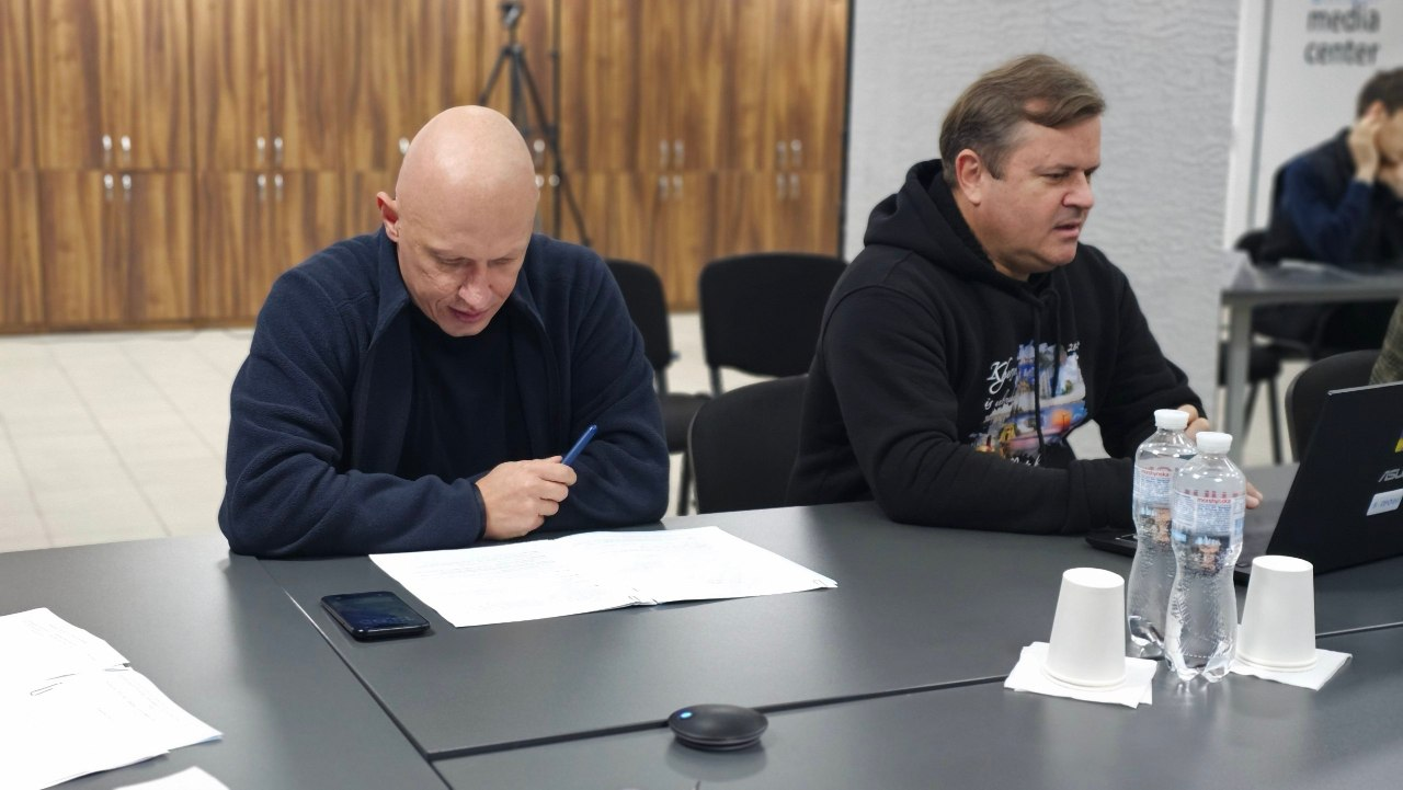За великим сірим столом зафіксовано заступників начальника Херсонської ОВА під час наради. Ліворуч сидітиме заступник начальника ХОВА Євгеній Ігнатенко у темно-синій кофті на блискавці, який схилився над робочими документами та тримає у правій руці синю ручку, а поруч на столі лежить його мобільний телефон. Праворуч від нього сидить заступник начальника ХОВА Володимир Клюцевський у чорному худі з яскравим малюнком, перед яким стоїть відкритий чорний ноутбук. На столі перед посадовцями також розставлено пластикові пляшки з водою та паперові склянки. На задньому плані приміщення видно зону з дерев'яними шафами, штативом, чорними офісними стільцями.