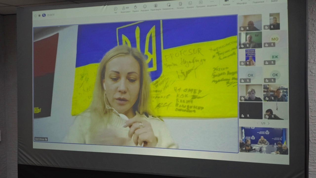На великому проєкційному екрані у залі засідань зафіксовано вікно онлайн-наради, яка проходить через відеозв'язок. Головну частину екрана займає трансляція спікерки — начальниці управління у справах ветеранів Херсонської ОДА Дар'ї Мазур. Білява жінка у світлому светрі з навушниками-вкладками у вухах виступає на тлі великого державного прапора України, розписаного патріотичними гаслами, автографами та побажаннями від військових (зокрема з написами «41 ОМБр», «Херсонщина»). Праворуч на екрані відображається вертикальна панель з іншими учасниками та вікнами відеозв'язку, серед яких у самому низу видно трансляцію із зали засідань Херсонської ОВА, де за великим сірим столом сидять керівники області.