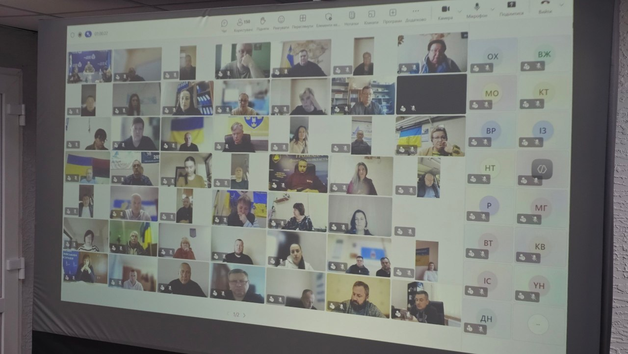 На великому проєкційному екрані у залі засідань зафіксовано загальне вікно онлайн-наради, яка проходить у форматі масштабної відеоконференції. Екран розділений на велику кількість невеликих квадратних вікон, де транслюються десятки підключених учасників наради — представників профільних структурних підрозділів ХОВА, районів та громад області, які беруть участь в обговоренні з робочих кабінетів або домашніх локацій. Деякі учасники виступають на тлі державних прапорів України, офіційних банерів чи робочих інтер'єрів. Праворуч на екрані відображається додаткова вертикальна панель із дволітерними іконками-ініціалами користувачів, які підключені до зустрічі без увімкнених відеокамер.
