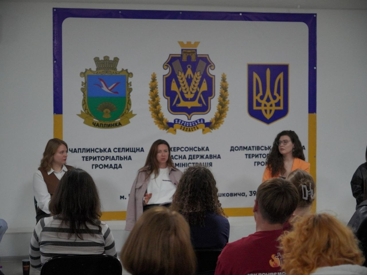 На світлині зафіксовано воркшоп з ментального здоров’я «Ментальне здоров’я: маленькі кроки до великої турботи» для переселенців з Херсонщини, який відбувся у координаційному центрі «Вільні разом» у Черкасах. У залі перед аудиторією на тлі великого білого банера з офіційними гербами Чаплинки, Херсонської області, Державним Гербом України та написами «Чаплинська селищна територіальна громада», «Херсонська обласна державна адміністрація» та «Долматівська сільська територіальна громада» сидять три спікерки заходу. Ліворуч сидить дівчина в білій блузі та коричневій жилетці, по центру — спікерка у білій кофті та розстебнутому світло-рожевому плащі, яка емоційно виступає та жестикулює руками, а праворуч — дівчина в окулярах та яскравому помаранчевому жакеті з пишним кучерявим волоссям. На передньому плані спиною до камери сидять слухачі та слухачки — внутрішньо переміщені особи з Херсонщини, які уважно слухають доповідь, зокрема видно жінку у смугастій кофті ліворуч та хлопця у бордовій футболці праворуч.