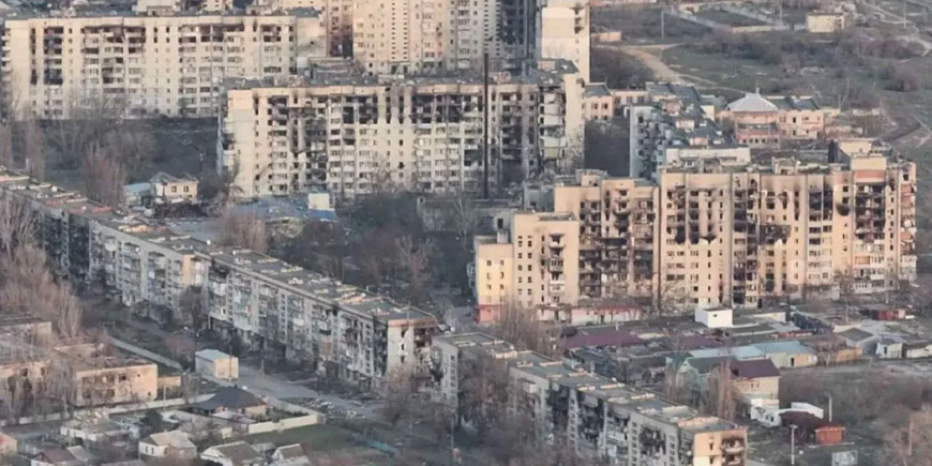 На світлині з висоти пташиного польоту показано панораму масштабних руйнувань житлових кварталів тимчасово окупованого міста Олешки на Херсонщині. У кадрі зафіксовано щільну міську забудову, яка зазнала катастрофічних пошкоджень внаслідок бойових дій та обстрілів. Вздовж вулиць розташовані багатоповерхові панельні будинки та хрущовки, у яких вибито вікна, повністю обпалені або зруйновані цілі секції, а стіни мають сліди численних влучань і кіптяви. На передньому плані та між висотками видніються залишки приватного сектору з понівеченими дахами невеликих одноповерхових хат, поодинокі вцілілі господарські споруди та рідкісні сухі осінні дерева. Весь міський пейзаж виглядає безлюдним і сірим під розсіяним денним світлом, що підкреслює масштаби гуманітарної катастрофи в регіоні.