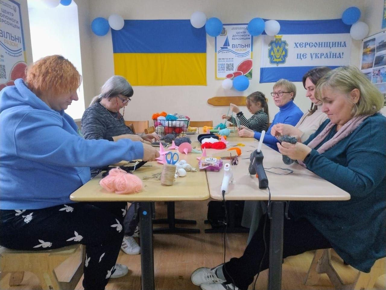 На світлині показано робочий процес переселенок із Херсонщини у кропивницькому координаційному центрі «Вільні разом» під час творчого майстер-класу. За двома зсунутими дерев'яними столами сидить група учасниць, які зосереджено виготовляють сувенірних відьом із ниток та декоративних матеріалів. Ліворуч на передньому плані сидить жінка з коротким рудим волоссям у блакитному худі та чорних штанях із принтом, яка зв'язує нитки, а поруч із нею працює жінка в сірому светрі та окулярах. На передньому плані праворуч зафіксована жінка у темно-бірюзовому кардигані та рожевому шарфі, яка обробляє деталі виробу, а ліворуч від неї сидить дівчина у світлому светрі. Далі по колу сидять жінка у синьому гольфі й окулярах та маленька дівчинка в смугастій кофті, яка займається рукоділлям під наглядом ще однієї учасниці. На столах розкладено різнокольорові мотки пряжі в кошику, ножиці, клей-пістолет, рожевий фетр, котушки шпагату та готові елементи сувенірів. На задньому плані кадру на світлій стіні, прикрашеній синьо-білими повітряними кульками, розвішано Державну символіку: ліворуч майорить Державний Прапор України, по центру розташований білий плакат із кавуном та написом «ЦЕНТР ДОПОМОГИ ХЕРСОНЦЯМ ВІЛЬНІ РАЗОМ», а праворуч височіє прапор Херсонської області із золотим гербом та патріотичним написом-гаслом «ХЕРСОНЩИНА ЦЕ УКРАЇНА!».