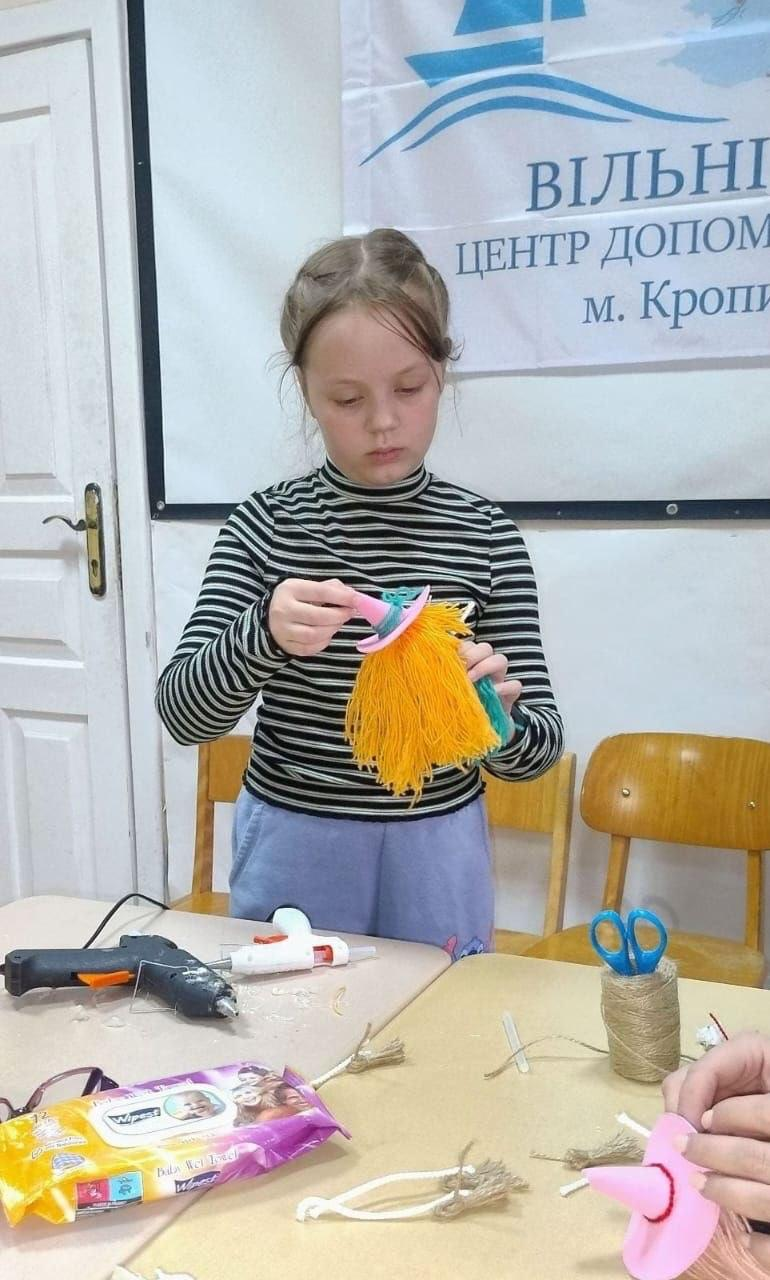 На світлині зображено маленьку херсонську переселенку під час створення сувенірної відьмочки на творчому майстер-класі в кропивницькому координаційному центрі «Вільні разом». Дівчинка у смугастому чорно-білому гольфі та бузкових штанях зосереджено стоїть біля столу, акуратно розправляючи та збираючи пальцями пишні яскраво-жовті нитки майбутнього виробу під рожевим капелюшком-конусом із фетру. Перед нею на робочому столі розкладено інструменти та матеріали для рукоділля: клей-пістолет, пачка вологих серветок, ножиці, моток шпагату, а праворуч у кадрі видно руки іншої учасниці, яка збирає аналогічний рожевий капелюшок. Позаду дитини розташовані дерев'яні стільці, білі міжкімнатні двері та височіє офіційний білий банер із синім фірмовим логотипом-корабликом та чіткими написом «ВІЛЬНІ РАЗОМ. ЦЕНТР ДОПОМОГИ ХЕРСОНЦЯМ м. Кропивницький».