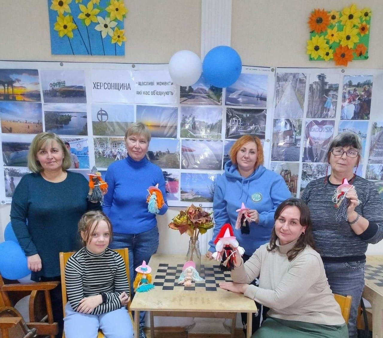 На світлині показано учасниць та організаторів творчого майстер-класу в кропивницькому координаційному центрі «Вільні разом», які радісно демонструють виготовлені власними руками сувенірні вироби. У кімнаті хабу біля невеликого столика з шахівницею, на якому стоїть ваза з осіннім листям та готові ляльки, зібралися п'ятеро жінок та маленька дівчинка. Ліворуч на стільці сидить дівчинка в чорно-білому смугастому гольфі та бузкових штанях, а праворуч від столу присіла жінка у світлому светрі та зеленій спідниці, яка тримає сувенірну відьмочку в червоно-чорному вбранні. У верхньому ряду зліва направо стоять: жінка у темно-бірюзовому кардигані; світловолоса жінка у синьому гольфі, яка тримає дві яскраві ляльки з жовтими та помаранчевими нитками; жінка з рудим волоссям у блакитному фірмовому худі з круглим логотипом; а також жінка в окулярах та сірій кофті, яка тримає виріб із рожевим капелюшком. Вся група позує на тлі великого білого стенда з фотовиставкою краєвидів рідного краю під загальною назвою «ХЕРСОНЩИНА — щасливі моменти які нас об'єднують», прикрашеного зверху синьо-білими кульками та яскравими дитячими аплікаціями з жовтими соняшниками й нарцисами.