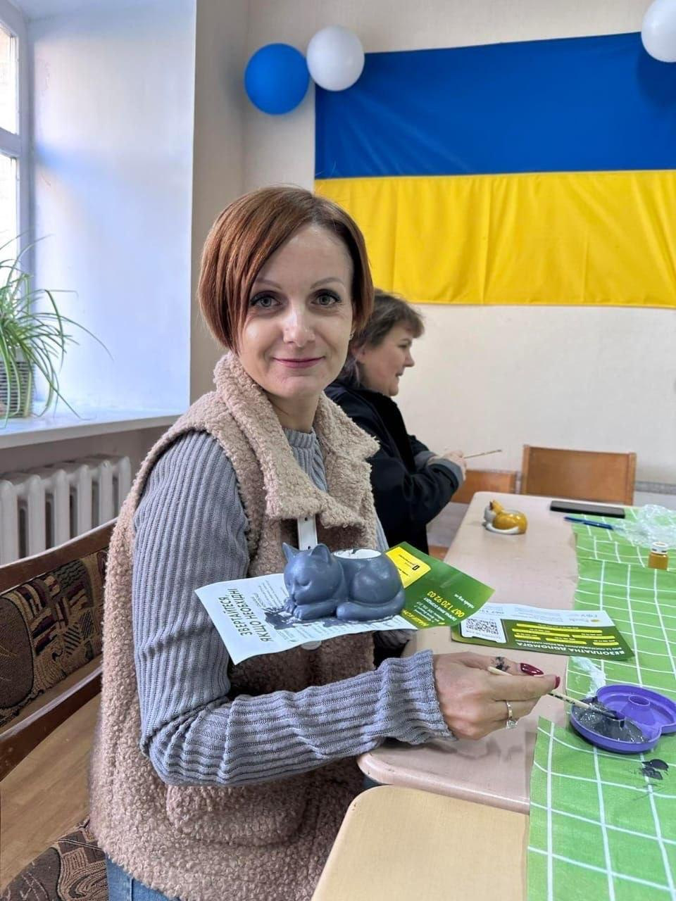 На світлині показано робочий момент арттерапевтичного майстер-класу з розмальовування гіпсових котів для переселенців із Херсонщини, який провела психологиня БФ «Рокада» Тетяна Мірошніченко у кропивницькому координаційному центрі «Вільні разом». На передньому плані за столом сидить усміхнена жінка-учасниця з короткою зачіскою у сірому светрі в рубчик та світлому хутряному жилеті, яка тримає перед собою вже розфарбовану фігурку сірого гіпсового котика. Під сувеніром лежить роздрукований інформаційний буклет БФ «Рокада» з великими написами «ЗВЕРТАЙТЕСЬ, ЯКЩО НЕОБХІДНО» та «БЕЗКОШТОВНА ДОПОМОГА». За нею в профіль зафіксовано іншу жінку у чорній куртці, яка зосереджено працює пензликом, а на столі ліворуч розкладено зелену скатертину в клітинку, палітру із фарбами та пензлі. На задньому плані кадру ліворуч розташоване велике вікно з кімнатною рослиною на підвіконні, а на стіні, прикрашеній синьо-білими повітряними кульками, майорить Державний Прапор України.
