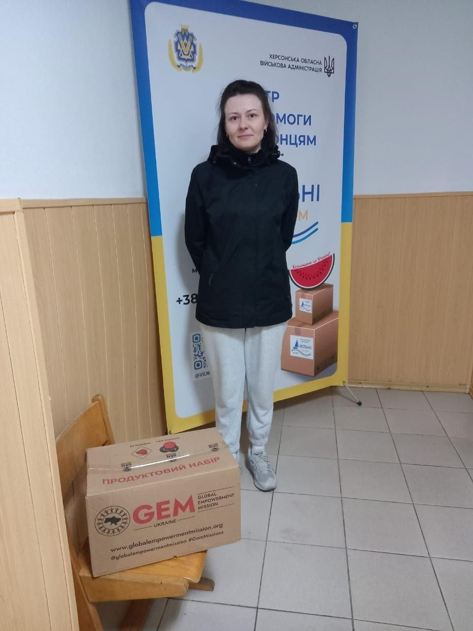 На світлині показано переселенку з Херсонщини під час видачі гуманітарної допомоги у кропивницькому координаційному центрі «Вільні разом». У коридорі хабу біля стіни, оббитої дерев'яною вагонкою, у повний зріст стоїть молода жінка з довгим темним волоссям, одягнена у чорну осінню куртку, світло-сірі спортивні штани та сірі кросівки. Ліворуч від неї на дерев'яному стільці встановлено велику картонну коробку гуманітарної допомоги з великим червоним написом «ПРОДУКТОВИЙ НАБІР», логотипом благодійної організації «GEM GLOBAL EMPOWERMENT MISSION UKRAINE» та адресою офіційного сайту. Позаду жінки височіє великий мобільний банер-ролап із гербом України, символікою Херсонщини та написами: «ХЕРСОНСЬКА ОБЛАСНА ВІЙСЬКОВА АДМІНІСТРАЦІЯ», «ЦЕНТР ДОПОМОГИ ХЕРСОНЦЯМ ВІЛЬНІ РАЗОМ», де також зображено малюнок часточки кавуна та коробки з допомогою.