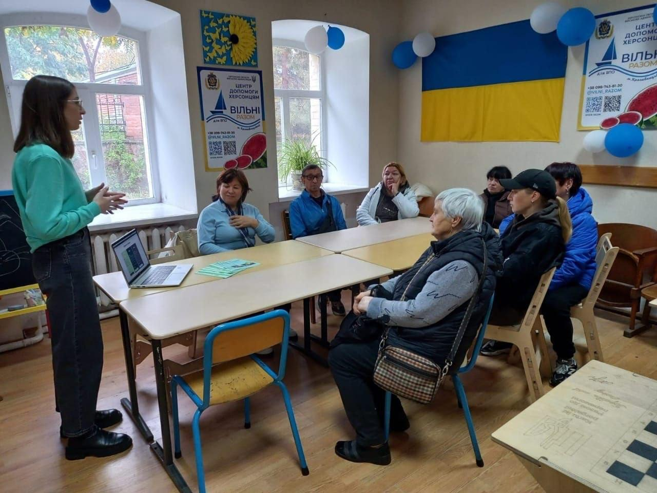 На світлині показано робочий момент заходу з фінансової грамотності для переселенців із Херсонщини, який провела керівниця відділення «ПриватБанку» Яна Максименко у кропивницькому координаційному центрі «Вільні разом». Ліворуч біля навчального столу у повний зріст стоїть молода спікерка в окулярах, одягнена в яскраво-зелений світшот та темні джинси, яка активно жестикулює й веде лекцію, презентуючи матеріали на відкритому сріблястому ноутбуці. За зсунутими дерев'яними столами півколом розмістилися слухачі — семеро людей старшого віку та молодь у верхньому одязі, які уважно слухають роз'яснення; зокрема на передньому плані праворуч сидить сивочола жінка в окулярах, сірій кофті з візерунками та темній жилетці з сумочкою через плече, а поруч із нею — дівчина у чорній кепці та куртці. На столі перед лекторкою розкладено зелені інформаційні буклети «ПриватБанку». Зустріч проходить у залі з паркетною підлогою на тлі великих вікон із кімнатними рослинами, де стіни прикрашені синьо-білими повітряними кульками, патріотичними плакатами із кавунами «ЦЕНТР ДОПОМОГИ ХЕРСОНЦЯМ ВІЛЬНІ РАЗОМ» та великим Державним Прапором України.