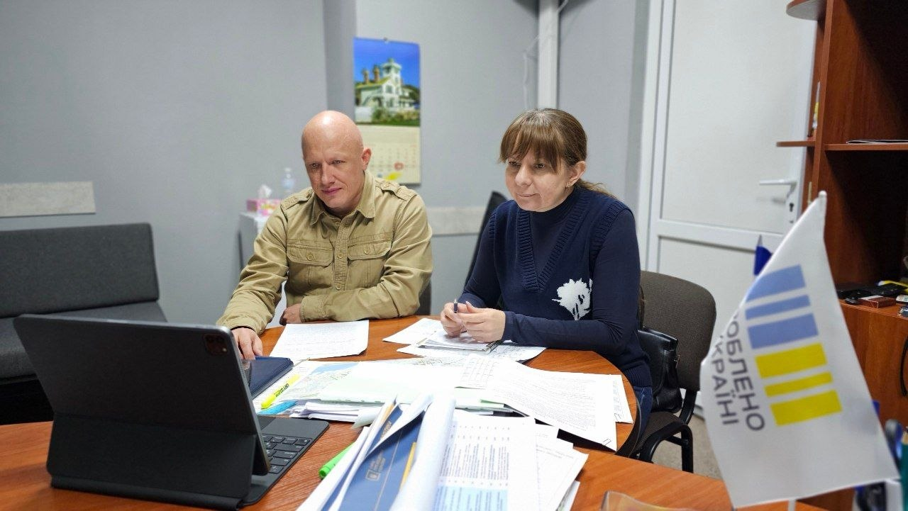 На світлині показано робочий момент онлайн-зустрічі заступника начальника Херсонської ОВА Євгенія Ігнатенка та директора Департаменту здоров'я ХОДА Тетяни Карчевич із головою НСЗУ Наталією Гусак щодо фінансування лікарень та Програми медичних гарантій. За круглим дерев'яним офіційним столом сидить керівництво області: ліворуч — Євгеній Ігнатенко в бежевій тактичній сорочці з накладними кишенями та погонами, а праворуч — Тетяна Карчевич у синій кофті з вишитим білим рослинним візерунком та зібраним у хвіст волоссям, яка тримає в руках синю ручку. Обоє зосереджено дивляться на екран відкритого чорного планшета-ноутбука на підставці, за допомогою якого здійснюється відеозв'язок. Робоча поверхня столу вщент заповнена роздрукованими паперами, офіційними звітами, картами та розгорнутими папками з документами, а на передньому плані праворуч встановлено настільний біло-синьо-жовтий прапорець із написом. Нарада проходить у кабінеті на тлі сірих стін, де позаду на стіні висить кольоровий настінний календар із зображенням будівлі, а праворуч помітно білі офісні двері та край дерев'яної шафи для документів.