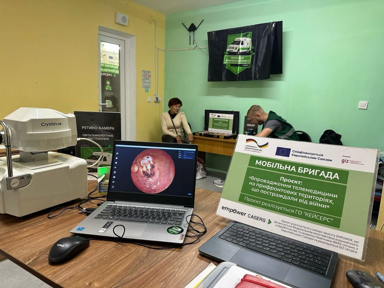 Медичний кабінет мобільної бригади ГО «CASERS», де на столі встановлено діагностичне обладнання для телемедицини: на передньому плані зліва розташована офтальмологічна ретино-камера Crystalvue, поруч із якою стоїть відкритий ноутбук із зображенням знімка очного дна пацієнта на екрані, а справа розміщено інформаційну табличку зеленого кольору з написами «Мобільна бригада» та описом проєкту «Впровадження телемедицини на прифронтових територіях, що постраждали від війни» від ГО «КЕЙСЕРС» (CASERS) за підтримки GIZ та Європейського Союзу, в той час як на задньому плані в кімнаті зі світло-зеленими стінами за іншим робочим місцем сидить чоловік-лікар, а поруч на стільці очікує жінка-пацієнт.