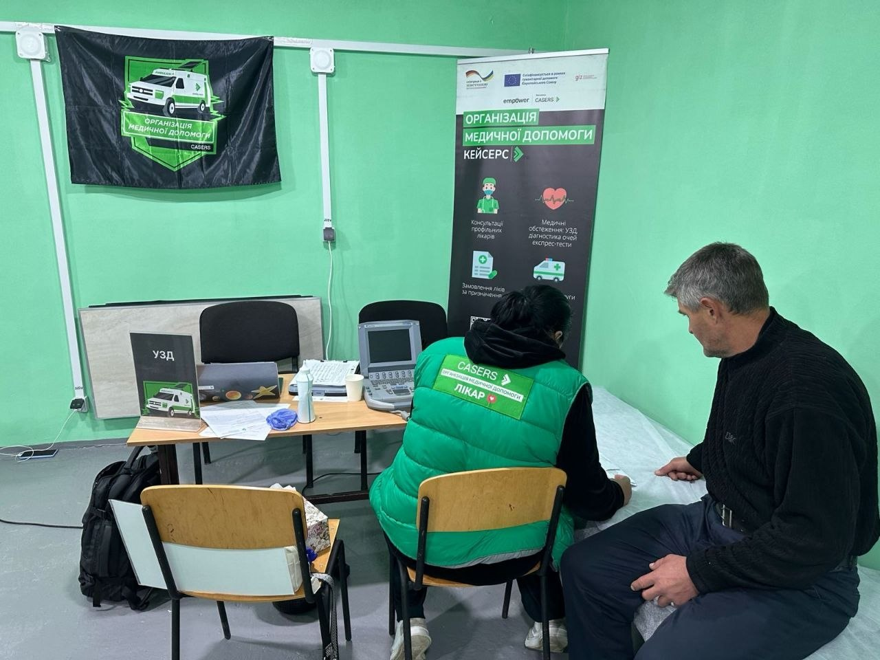 Процес консультації в кабінеті ультразвукової діагностики під час виїзду мобільної бригади ГО «CASERS»: у кімнаті зі світло-зеленими стінами по центру спиною до камери сидить лікар у яскраво-зеленому жилеті з написом «CASERS ЛІКАР», яка заповнює медичні документи, а праворуч на кушетці сиділа в профіль людина — сивочолий пацієнт-чоловік у чорній кофті, який слухає спеціаліста, при цьому зліва розташований робочий стіл із портативним апаратом УЗД, табличкою «УЗД» та принтером, а на задньому плані видно великий темний банер-ролап із написом «Організація медичної допомоги КЕЙСЕРС» та чорний прапор організації на стіні із зображенням медичного автомобіля.