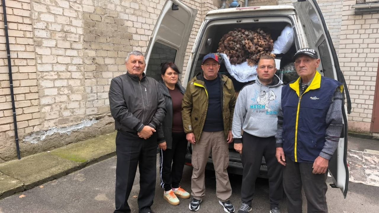 Волонтерська допомога військовим від громад та благодійників для Сил оборони: на фоні старої світло-цегляної стіни будівлі біля повністю відчиненого заднього вантажного відсіку білого мікроавтобуса, в якому видно складені коробки, пакунки та велику згорнуту коричневу маскувальну сітку, стоять у ряд п'ятеро людей у цивільному одязі, де по центру в коричнево-зеленій куртці та синій кепці стоїть начальник Чулаківської СВА Руслан Русаль, ліворуч від нього стоять чоловік у чорній шкіряній куртці та жінка у темній кофті й кросівках із помаранчевими шнурками, а праворуч — молодий чоловік у сірому худі з написом та літній чоловік у синьому жилеті з жовтим коміром і чорній кепці, які разом організували збір та доставку допомоги.