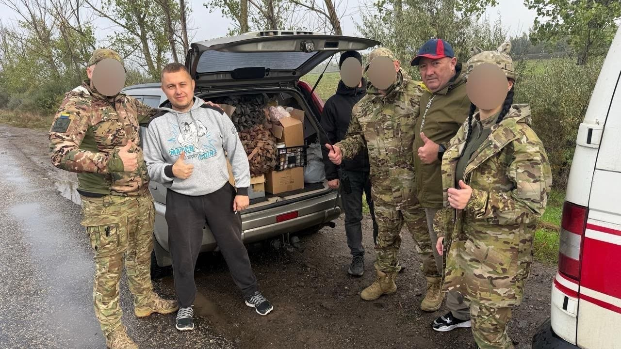 Передача волонтерської допомоги українським військовим безпосередньо на місці дислокації підрозділу: на ґрунтовій дорозі біля поля стоїть сірий позашляховик із відчиненим багажником, завантаженим картонними коробками з продуктами та згорнутими коричневими маскувальними сітками, а поруч зібралася група з шести людей, причому обличчя чотирьох із них приховані з міркувань безпеки за допомогою цифрового розмиття — серед присутніх начальник Чулаківської СВА Руслан Русаль у коричнево-зеленій куртці та синьо-червоній кепці (стоїть другим праворуч), чоловік у сірому спортивному худі, троє військовослужбовців ЗСУ у камуфляжній формі «мультикам» (зокрема 21-річна захисниця, донька загиблого Героя громади, яка стоїть на передньому плані праворуч у військовій кепці) та чоловік у чорній куртці з капюшоном, при цьому більшість учасників зустрічі підіймають великий палець угору.