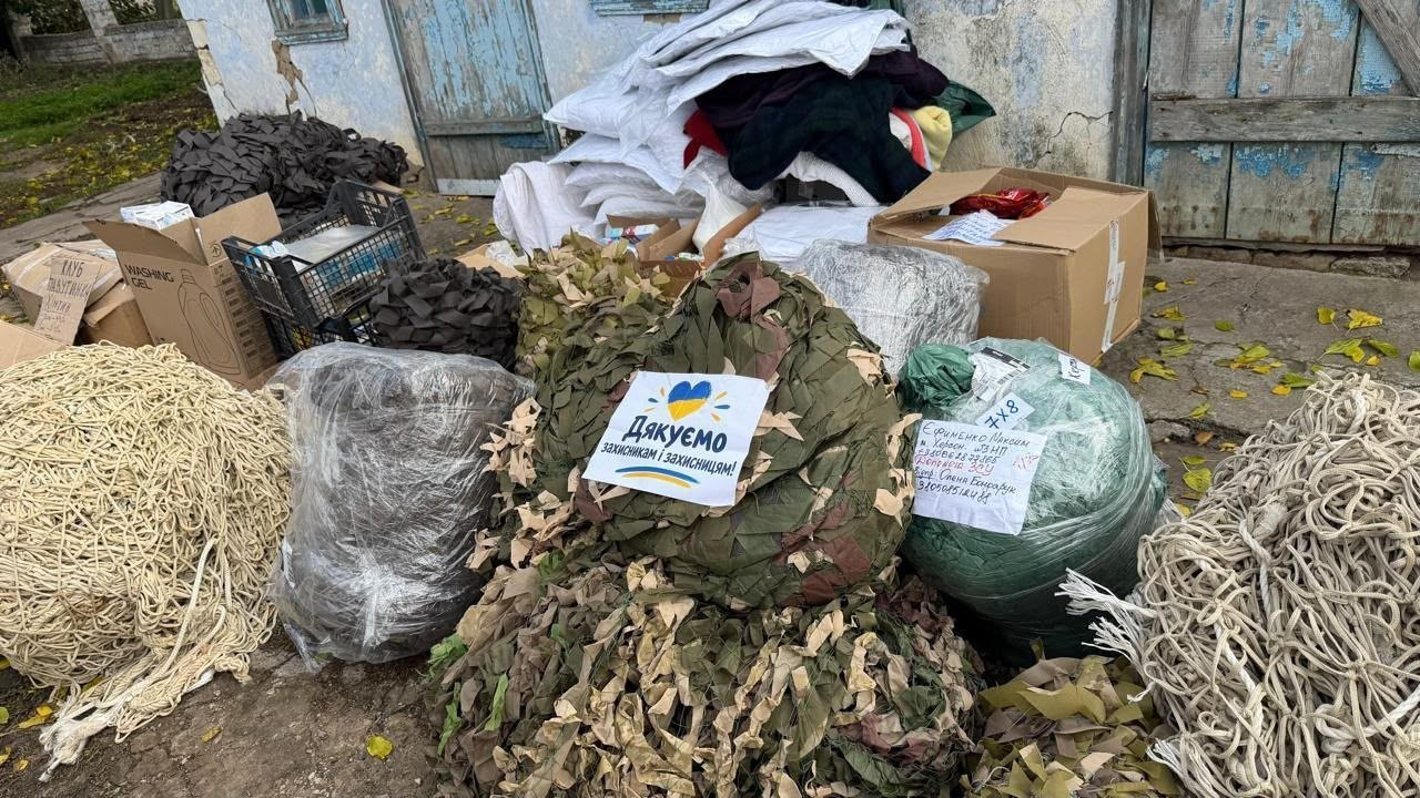Гуманітарна допомога та маскувальні сітки, підготовлені для відправки Силам оборони від Чулаківської громади та волонтерів: на подвір'ї біля старої будівлі з блакитними дерев'яними дверима складено велику купу речей, серед яких на передньому плані виділяються кілька великих рулонів маскувальних сіток різних відтінків — білі, зелені та коричневі, а по центру на одній із сіток прикріплено папірець із малюнком синьо-жовтого серця та написом «Дякуємо захисникам і захисницям!», у той час як на задньому плані розміщені картонні коробки, пластикові ящики з допомогою та велика стопка ковдр або теплих речей у білих мішках.