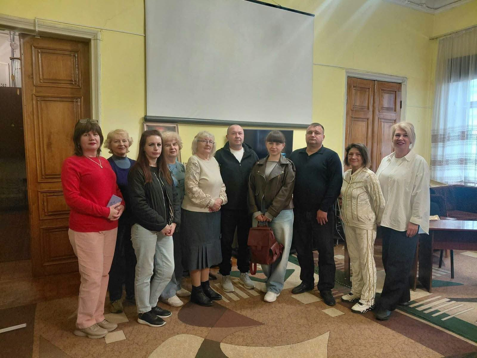 Учасники творчої зустрічі «Сергій Шаповалов — співець вічної краси» у відділі мистецтв Кіровоградської обласної бібліотеки для юнацтва ім. Є. Маланюка: у залі зі світло-жовтими стінами, високими дерев'яними дверима та великим білим проєкційним екраном на стіні стоїть група людей, серед яких фахівці координаційного центру «Вільні разом» та краєзнавиця Олена Классова; на передньому плані зліва виділяється жінка в яскраво-червоній кофті та рожевих штанах, а праворуч — жінка в білій блузі та чорних штанах; усі присутні вишикувалися в один ряд на коричнево-бежевому візерунчастому килимі та посміхаються в камеру для спільного пам'ятного фото.