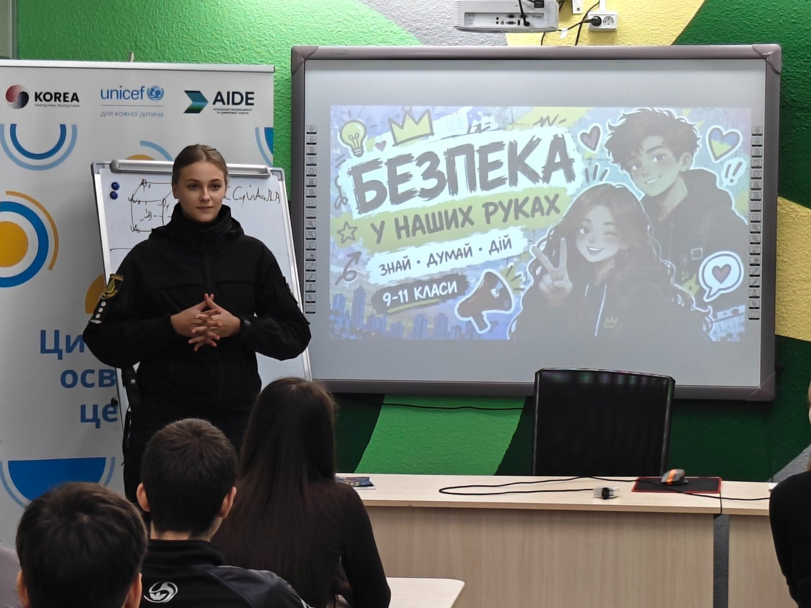 Проведення уроку безпеки для старшокласників Високопільського ліцею в сучасному цифровому освітньому центрі: ліворуч біля великого мультимедійного екрана стоїть молода інспекторка ювенальної превенції у чорній формі поліції із зібраним у пучок волоссям, яка тримає руки перед собою та спілкується з учнями, демонструючи головний слайд презентації на тему «БЕЗПЕКА У НАШИХ РУКАХ. ЗНАЙ • ДУМАЙ • ДІЙ. 9-11 КЛАСИ» із мальованими зображеннями підлітків, у той час як на передньому плані спиною до камери сидять за партами та слухають лекцію хлопець у чорній спортивній кофті з білим круглим логотипом та дівчина з довгим темним волоссям, а зліва за поліцейською помітно білий банер із логотипами KOREA, unicef та AIDE й написом «Цифровий освітній центр», при цьому на задньому плані під екраном облаштовано великий світлий робочий стіл із чорним офісним кріслом та комп'ютерною мишкою на килимку.