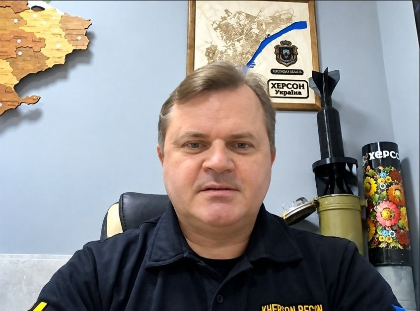 Заступник голови Херсонської ОДА Володимир Клюцевський під час онлайн-засідання гуманітарного штабу: по центру крупним планом зображено чоловіка середнього віку зі світло-русявим волоссям, який одягнений у чорну брендовану сорочку-поло з жовтим написом «KHERSON REGION» на грудях та синьо-жовтим прапором на правому рукаві; він сидить в офісному кріслі на тлі світло-блакитної стіни кабінету, де ліворуч закріплено велику дерев'яну декоративну карту України, а безпосередньо над його головою висить рамка з картою-схемою річки Дніпро, гербом Херсонської області та написом «ХЕРСОН Україна»; праворуч на робочому столі зафіксовано військові сувеніри-експонати — хвостовик від мінометної міни, гільзу від снаряда артилерії та розписаний яскравими традиційними квітами чорний тубус від гранатомета з написом «ХЕРСОН».