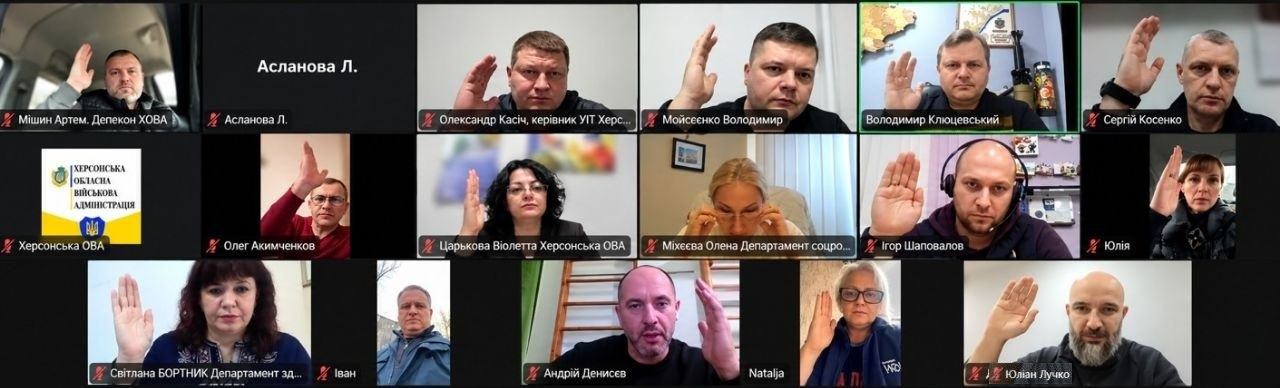 Скриншот онлайн-засідання гуманітарного штабу при Херсонській ОВА під керівництвом заступника голови Херсонської ОДА Володимира Клюцевського: загальний екран відеоконференції розділений на шістнадцять вікон із учасниками зустрічі, більшість із яких підняли праву руку на знак одностайного голосування за рішення штабу. У верхньому ряду праворуч виділяється вікно керівника засідання Володимира Клюцевського, який сидить у робочому кабінеті на тлі військових експонатів та карти України. Серед присутніх на засіданні присутні керівники профільних структурних підрозділів ОДА: директор Департаменту розвитку економіки Артем Мішин (верхнє ліве вікно), начальник управління інформаційних технологій Олександр Касіч (верхній ряд, третій ліворуч), начальник юридичного відділу Володимир Мойсеєнко (верхній ряд, четвертий ліворуч), директорка Департаменту реалізації гуманітарної політики Віолета Царькова (другий ряд, третя ліворуч), директорка Департаменту соціального розвитку Олена Міхєєва (другий ряд, по центру) та начальник управління з питань децентралізації та розвитку громад Юліан Лучко (нижнє праве вікно). Інші вікна конференції містять зображення решти учасників, серед яких Сергій Косенко, Олег Акимченков, Ігор Шаповалов, Юлія, Світлана Бортник, Іван, Андрій Денисов та Наталя, у той час як вікно Л. Асланової залишається з вимкненою камерою на чорному тлі.