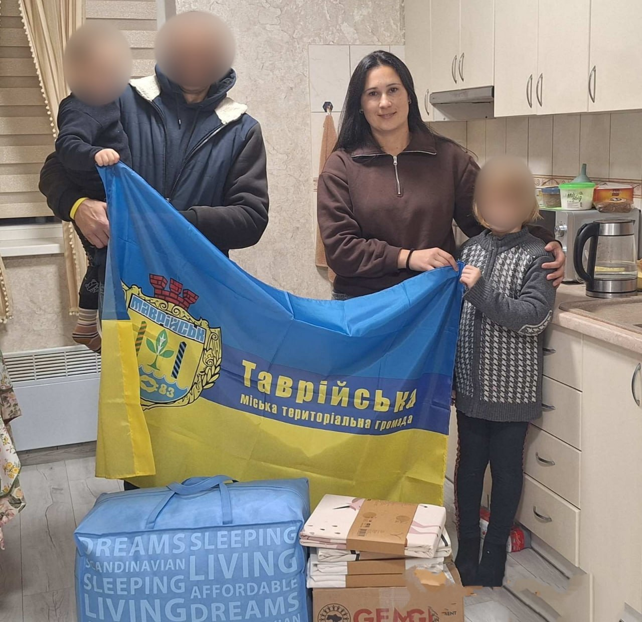 Зустріч начальниці служби у справах дітей Таврійської міської ради з родиною переселенців у їхньому тимчасовому помешканні: у світлій кухонній кімнаті біля стіни та кухонних шаф стоїть група людей, які разом розгорнули та тримають перед собою великий синьо-жовтий прапор із гербом міста Таврійськ та написом «Таврійська міська територіальна громада»; по центру стоїть начальниця служби у справах дітей у коричневій кофті на блискавці, яка посміхається в камеру; ліворуч від неї стоїть чоловік у темній зимовій куртці з хутряним коміром, який тримає на руках маленьку дитину, а праворуч — дівчинка у сірому в'язаному светрі та темних штанах, при цьому обличчя чоловіка, маленької дитини та дівчинки приховані цифровим розмиттям з міркувань безпеки; на передньому плані на підлозі перед ними стоять подарунки — велика синя прямокутна сумка з постільною білизною та написами «JYSK SCANDINAVIAN LIVING AFFORDABLE DREAMS SLEEPING», кілька картонних коробок і пакунків, а на задньому плані праворуч помітно частину кухонної стільниці з чайником, мікрохвильовою піччю та побутовими речами.
