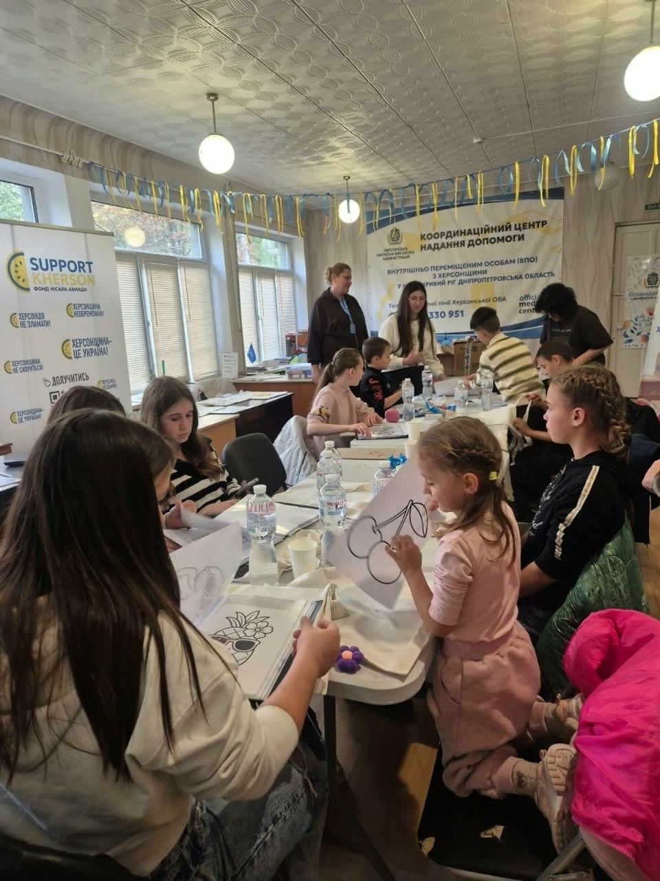 Проведення творчого майстер-класу «Яскраві емоції осені» від БФ «Рокада» для юних переселенців у Криворізькому координаційному центрі «Вільні разом»: у великій світлій залі з вікнами, завішаними жалюзі, та круглими лампами під стелею за довгими білими столами сидить велика група дітей різного віку, які зосереджено займаються малюванням та творчістю; на передньому плані зліва дівчинка з довгим темним волоссям розглядає паперовий альбом із контурами малюнків, поруч із нею сидить дівчина у смугастій кофті, а праворуч на колінах на стільці піднялася маленька дівчинка із заплетеним волоссям у рожевому костюмі, яка тримає в руках аркуш із намальованими вишнями; посеред столу розставлено пляшки з водою, пластикові стаканчики та олівці, а вздовж верхньої частини стін розвішано жовто-блакитні паперові гірлянди; на задньому плані ліворуч встановлено білий інформаційний стенд благодійного фонду «SUPPORT KHERSON» з написами «ХЕРСОНЦІ НЕЗЛАМНІ!» та «ХЕРСОНЩИНА — ЦЕ УКРАЇНА!», а по центру на стіні висить великий банер «КООРДИНАЦІЙНИЙ ЦЕНТР НАДАННЯ ДОПОМОГИ ВНУТРІШНЬО ПЕРЕМІЩЕНИМ ОСОБАМ (ВПО) З ХЕРСОНЩИНИ У М. КРИВИЙ РІГ ДНІПРОПЕТРОВСЬКА ОБЛАСТЬ», біля якого стоять та допомагають дітям представниці фонду й організатори.