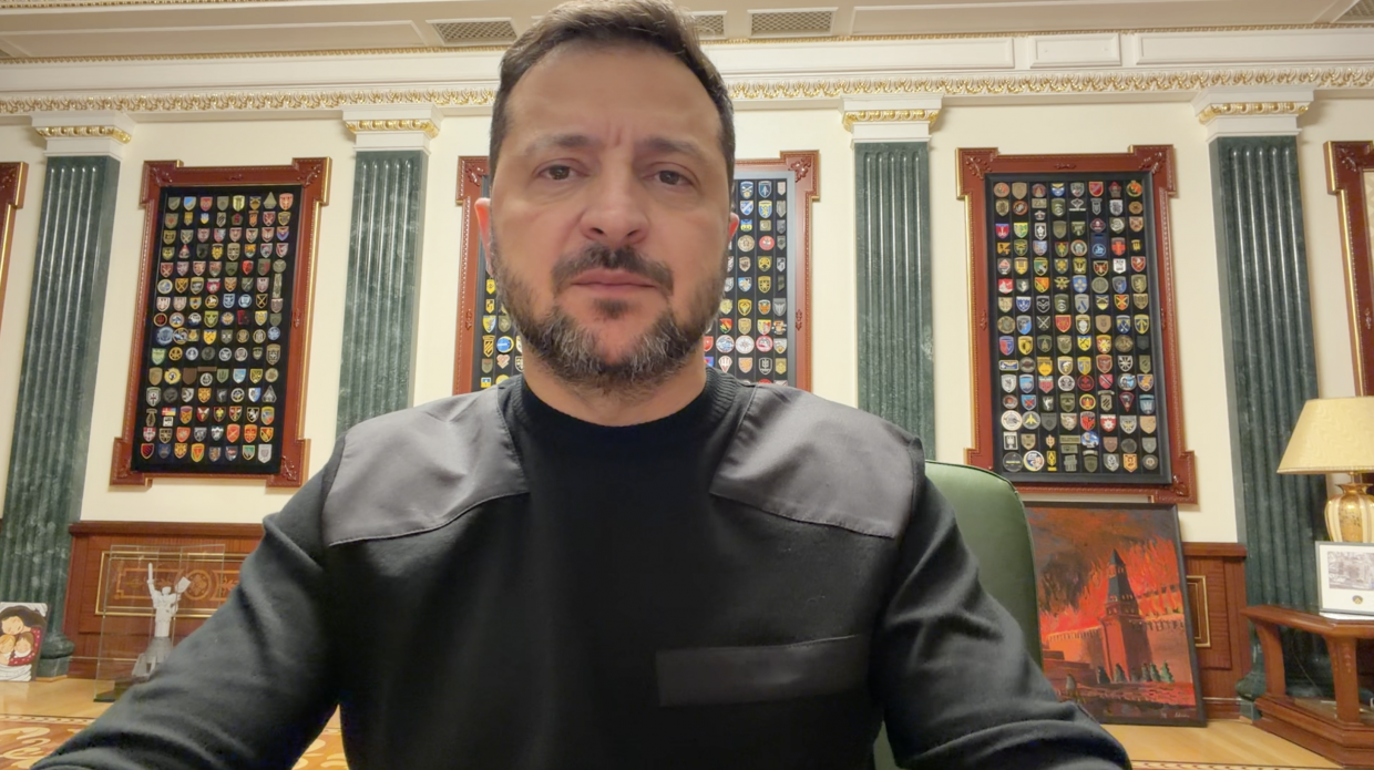 Офіційне відеозвернення Президента України Володимира Зеленського щодо військової операції «Велес» на Куп’янському напрямку: по центру великим планом зображено Президента у чорному світшоті, який сидить у зеленому шкіряному кріслі та говорить у камеру; на задньому плані в кабінеті вздовж світлої стіни з темно-зеленими мармуровими колонами та золотим карнизом висять у масивних рамах чотири великі колекційні стенди з сотнями різнокольорових військових шевронів Сил оборони; праворуч на підлозі стоїть картина в рамі із зображенням палаючого Кремля, а в кутку помітно частину дерев'яного столика з настільною лампою.
