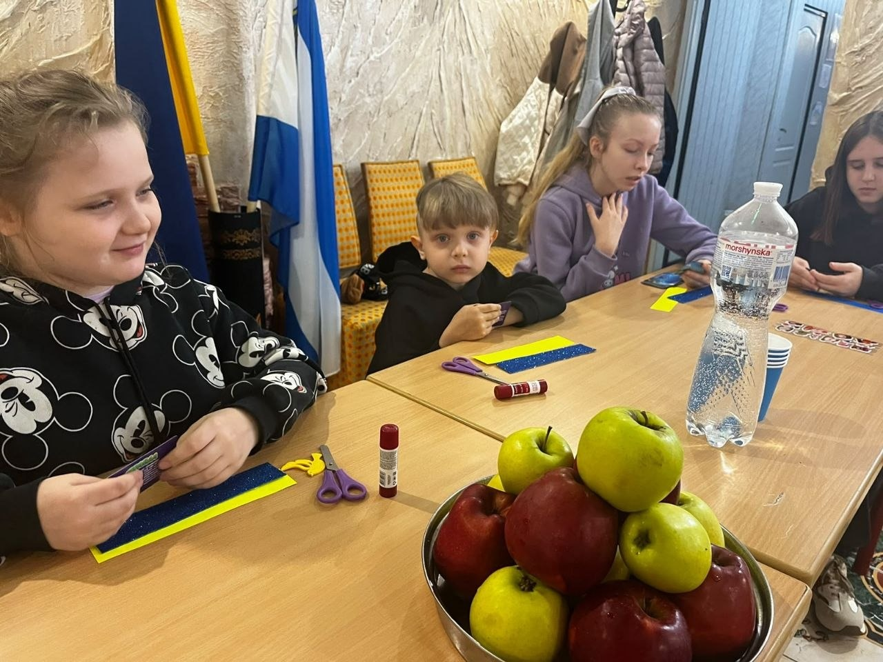 Діти-переселенці з Херсонщини під час творчого майстер-класу в Одеському координаційному центрі «Вільні разом»: за довгим дерев'яним столом сидить група дітей, які займаються виготовленням паперових поробок та закладок для книг; безпосередньо на передньому плані ліворуч усміхається дівчинка в чорній кофті з малюнками Міккі Мауса, перед якою на столі лежать ножиці, клей-олівець та синьо-жовта паперова основа; по центру столу стоїть велика металева миска, наповнена стиглими червоними та зеленими яблуками, а поруч — велика пластикова пляшка мінеральної води «Моршинська»; за столом далі сидять маленький хлопчик у чорному худі, дівчина у бузковій кофті з резинкою-бантом на волоссі та ще одна дівчинка праворуч, яка розглядає кольорові картки; на задньому плані ліворуч у кутку кімнати встановлено на стійках державний прапор України та прапор Херсонської області, а вздовж стіни стоять м'які стільці з помаранчевою оббивкою та висить верхній одяг.