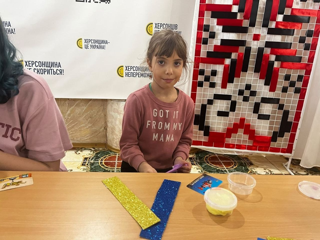 Маленька учасниця творчого заходу «Рідна мова єднає» під час майстер-класу в Одеському координаційному центрі «Вільні разом»: по центру за дерев'яним столом сидить дівчинка з челкою та зібраним волоссям у рожевому світшоті з білим текстовим принтом «GOT IT FROM MY MAMA», яка посміхається та дивиться в камеру, тримаючи в руках матеріали для поробки; на столі перед нею викладено блискучі заготовки для паперової синьо-жовтої закладки у формі галочки, кольорову ігрову картку та круглу прозору баночку з пластиліном або масою для ліплення; на задньому плані ліворуч стоїть білий інформаційний стенд із патріотичними гаслами «ХЕРСОНЩИНА НЕПЕРЕМОЖНА!», «ХЕРСОНЩИНА — ЦЕ УКРАЇНА!» та «ХЕРСОНЩИНА СКОРИТЬСЯ!», а праворуч облаштовано велике декоративне панно на стійці з традиційним українським геометричним вишитим орнаментом у червоно-чорних та білих кольорах.