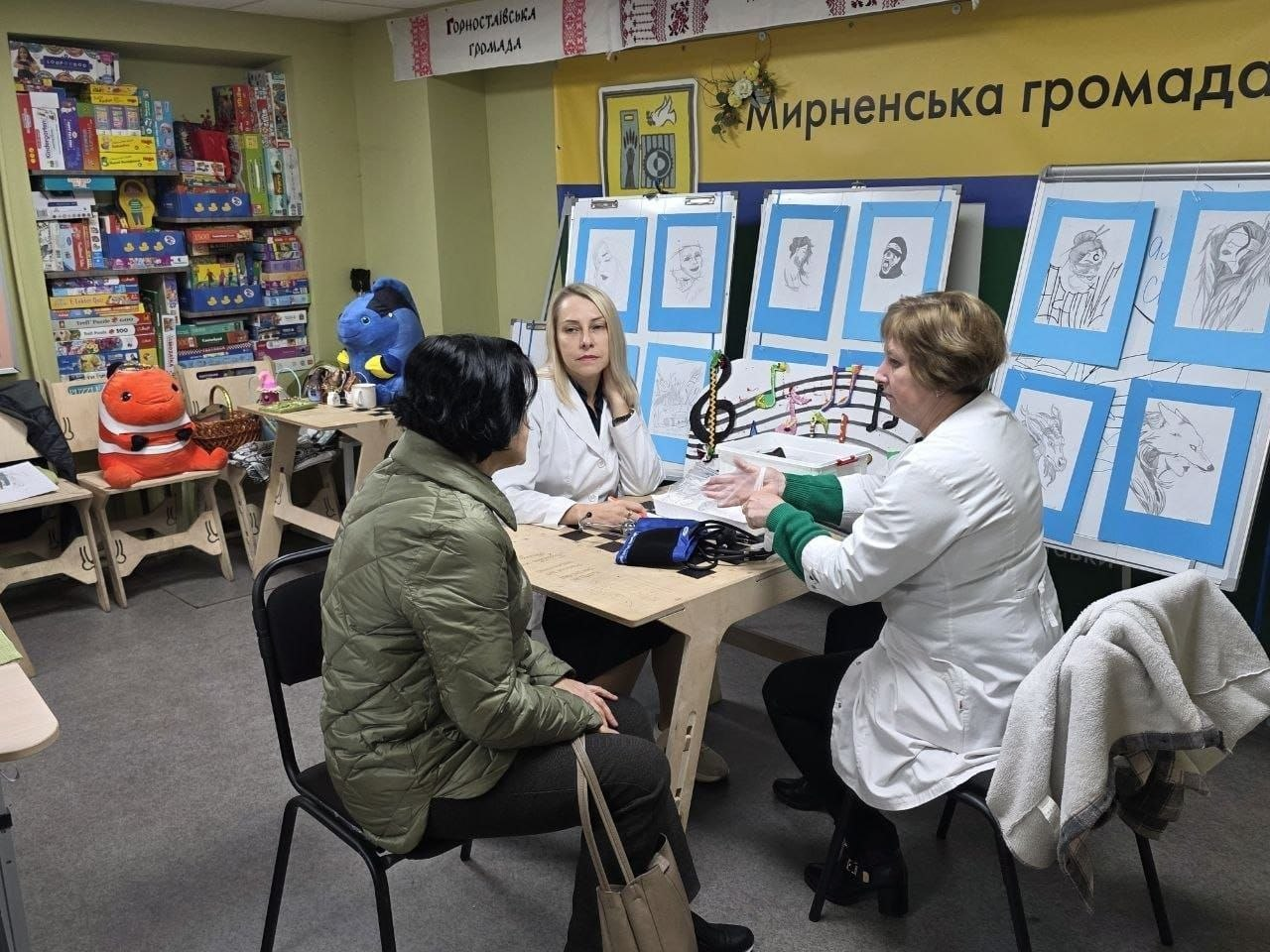 Прийом пацієнтів-переселенців із Херсонщини сімейними лікарями у Дніпровському координаційному центрі «Вільні разом»: у кабінеті зі світло-зеленими стінами за кутовим дерев'яним столом сидять три жінки під час медичної консультації; на передньому плані зліва спиною до камери на чорному стільці сиділа відвідувачка у зеленій куртці та темних штанах із бежевою сумкою на плечі; навпроти неї по центру столу сидить білява жінка-лікар у білому медичному халаті, яка підперла голову рукою та уважно слухає колегу; праворуч боком до камери сидить інша лікарка у білому халаті та зеленому светрі під ним, яка емоційно жестикулює руками, роз'яснюючи інформацію; на столі між ними лежать медичні картки, бланки та чорний тонометр для вимірювання артеріального тиску; на задньому плані кабінету ліворуч стоять полиці, повністю заставлені різнокольоровими коробками з дитячими настільними іграми, а на стільцях поруч лежать великі м'які іграшки; стіни кімнати прикрашені офіційними банерами з написами «Горностаївська громада» та великим жовто-синім стендом «Мирненська громада», під якими на дошках та мольбертах виставлено численні дитячі малюнки й портрети, оформлені у блакитні паспарту, а також макет музичного нотного стану з кольоровими нотами.