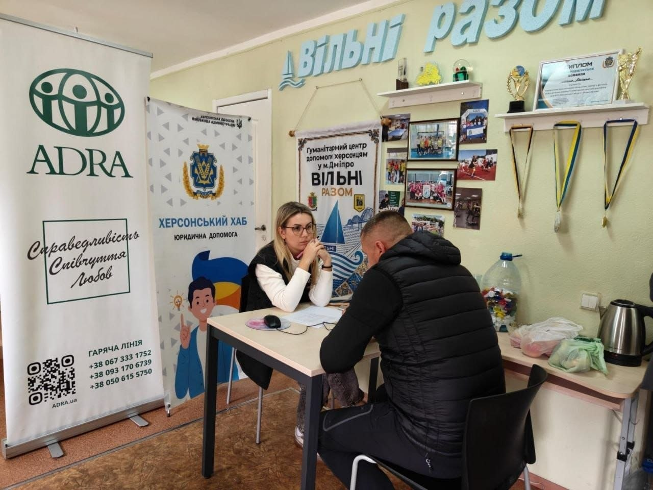 Надання індивідуальних юридичних консультацій для переселенців із Херсонщини в Дніпровському координаційному центрі «Вільні разом»: у світлому кабінеті за прямокутним столом сидять двоє людей під час правової консультації; ліворуч обличчям до камери сидить жінка-юрисконсульт в окулярах та світлій кофті, яка уважно слухає відвідувача, склавши пальці біля обличчя; навпроти неї на передньому плані праворуч спиною до камери на чорному стільці сидить чоловік-переселенець у чорному стьобаному жилеті з капюшоном; на столі між ними лежать робочі папери, бланки та комп'ютерна мишка; з лівого боку встановлено великі рекламні банери: перший — благодійної організації «ADRA» з гаслами «Справедливість. Співчуття. Любов» та контактами гарячої лінії, другий — «ХЕРСОНСЬКИЙ ХАБ. ЮРИДИЧНА ДОПОМОГА» з офіційним гербом; на задньому плані на світло-зеленій стіні закріплено великий напис «Вільні разом», під яким висить інформаційний стенд «Гуманітарний центр допомоги херсонцям у м. Дніпро ВІЛЬНІ РАЗОМ» із фотографіями заходів, а праворуч на полиці виставлено спортивні кубки, диплом та медалі на стрічках.