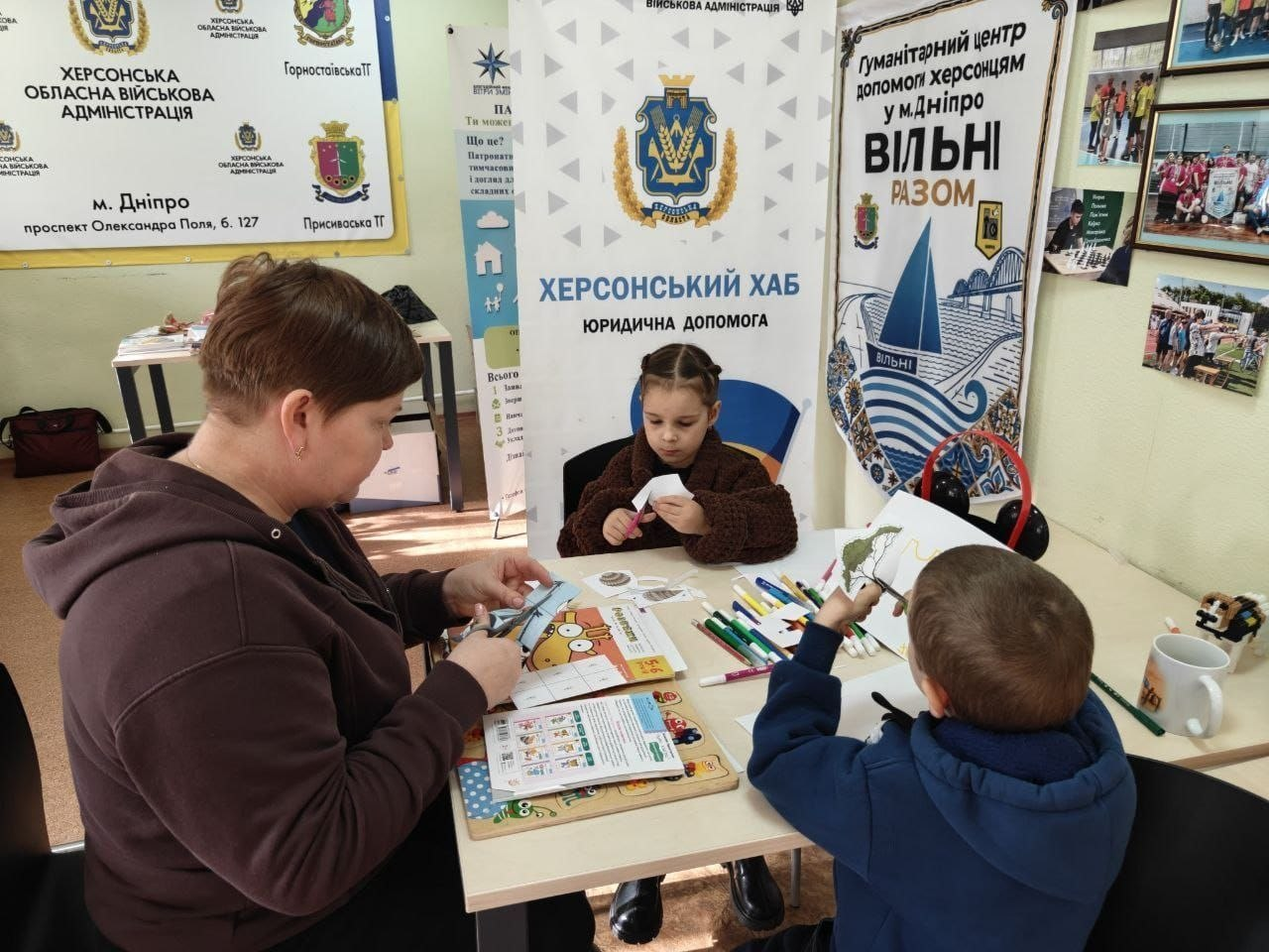 Заняття дитячого гуртка «Мама + Я» для родин переселенців із Херсонщини в Дніпровському координаційному центрі «Вільні разом»: за світлим дитячим столом сидить група людей, які займаються творчістю та розвивальними іграми; на передньому плані ліворуч боком до камери сидить мама в темній кофті із зібраним волоссям, яка допомагає дітям, а праворуч спиною до камери сидить маленький хлопчик у синій кофті з капюшоном, який тримає в руці кольоровий олівець; навпроти них по центру за столом сидить дівчинка в коричневому пухнастому светрі, яка зосереджено вирізає щось із паперу ножицями; на столі перед ними розкладено фломастери, дитячу книжку із зображенням зеленого монстрика Ам Няма та рудого пухнастого персонажа Тос, розвивальну дерев'яну дошку-вкладку з цифрами та бланки із завданнями; кімната хабу повністю завішана офіційними стендами: ліворуч стоїть великий жовтий банер «ХЕРСОНСЬКА ОБЛАСНА ВІЙСЬКОВА АДМІНІСТРАЦІЯ м. Дніпро» з гербами Горностаївської та Присиваської ТГ, по центру — білий стенд «ХЕРСОНСЬКИЙ ХАБ. ЮРИДИЧНА ДОПОМОГА», а праворуч на стіні закріплено інформаційний плакат «Гуманітарний центр допомоги херсонцям у м. Дніпро ВІЛЬНІ РАЗОМ» із фотографіями дитячих заходів.