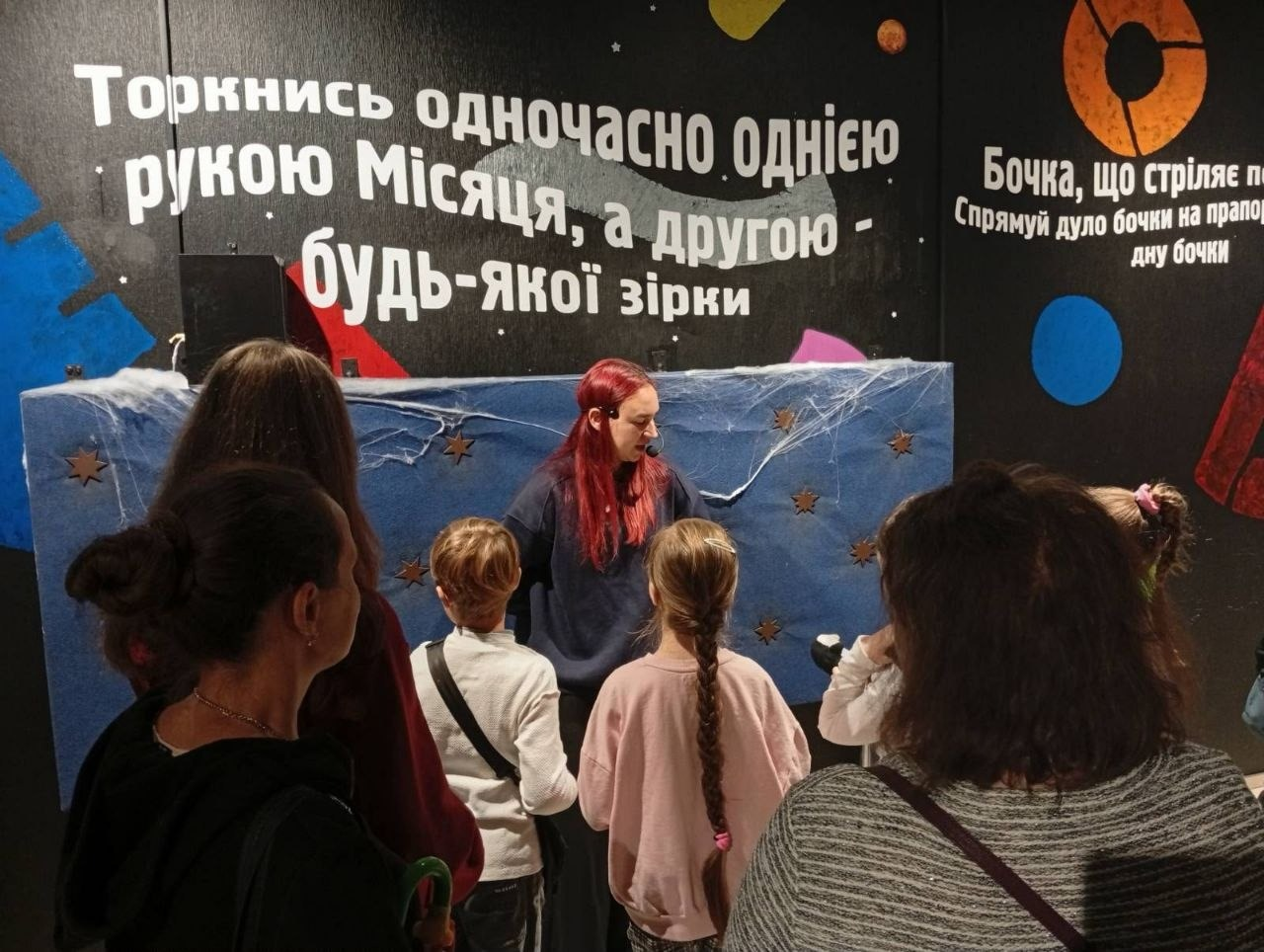 Діти-переселенці з Херсонщини разом із батьками під час інтерактивної екскурсії в Музеї науки в Одесі: у темній залі біля експоната з космічною тематикою стоїть група екскурсантів — переважно жінки та діти шкільного віку в повсякденному осінньому одязі, які повернуті спиною до камери й уважно слухають екскурсовода. Перед ними за синьою декоративною перегородкою, прикрашеною штучною білою павутиною та маленькими зірочками, стоїть дівчина-гід з яскравим довгим червоно-рудим волоссям та мікрофоном-гарнітурою, одягнена в темний світшот. Над її головою на чорній стіні нанесено великий білий напис-інструкцію: «Торкнись одночасно однією рукою Місяця, а другою - будь-якої зірки», а праворуч на стіні помітно частину іншого інструктивного тексту: «Бочка, що стріляє п... Спрямуй дуло бочки на прапо... дну бочки» поруч із кольоровими геометричними фігурами.