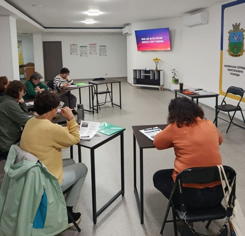Заняття з англійської мови для переселенців із Херсонщини в Черкаському координаційному центрі «Вільні разом»: у просторій світлій залі з білими стінами за окремими чорними металевими столами сидить група жінок середнього та старшого віку в повсякденному одязі (кофтах, светрах), які зосереджено виконують навчальні вправи та роблять записи у зошитах і на роздрукованих аркушах. Усі учасниці повернуті спиною, боком або напівпрофілем до камери, повністю занурені в навчальний процес. На задньому плані по центру на стіні встановлено великий плазмовий екран з англійським текстом презентації на яскравому червоному тлі, під яким стоїть світла тумба з вазою жовтих квітів, а праворуч на стіні облаштовано великий офіційний жовто-синій стенд із гербом та написом «ЧАПЛИНСЬКА СЕЛИЩНА ТЕРИТОРІАЛЬНА ГРОМАДА».