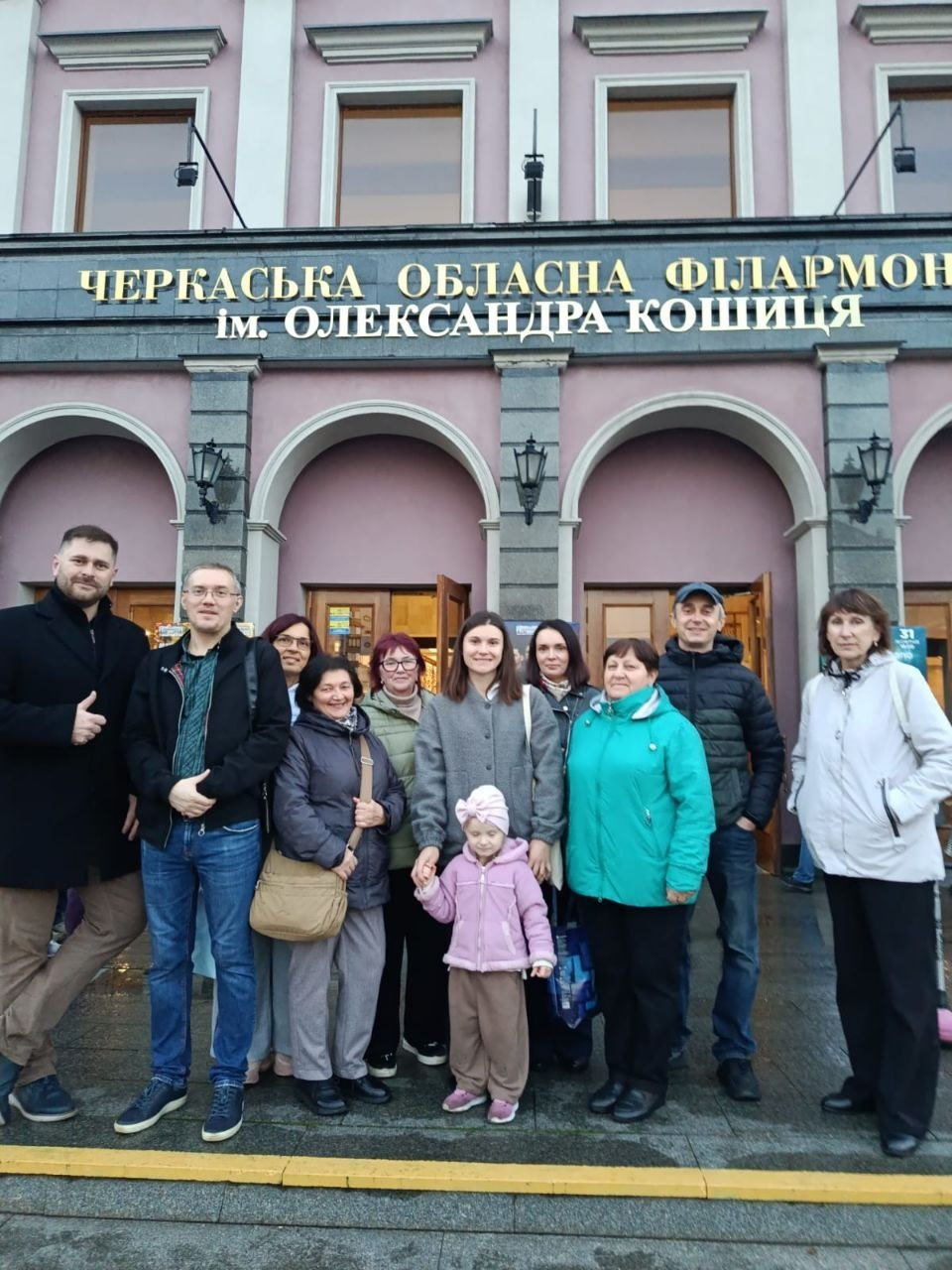 Група переселенців із Херсонщини перед будівлею Черкаської обласної філармонії імені Олександра Кошиця після відвідування концерту симфонічного оркестру: на тлі рожево-сірого класичного фасаду філармонії з арковими входами, колонами та старовинними ліхтарями стоїть група людей, які прийшли на культурний захід за сприяння координаційного центру «Вільні разом». Учасники зустрічі — чоловіки, жінки різного віку та маленька дівчинка в рожевій куртці й шапці по центру — одягнені в теплий осінній одяг, куртки та джинси. Усі присутні вишикувалися в один ряд на тротуарі перед вхідними дверима закладу, посміхаються та разом позують для пам'ятного групового знімка під великим золотим написом на фасаді: «ЧЕРКАСЬКА ОБЛАСНА ФІЛАРМОНІЯ ім. ОЛЕКСАНДРА КОШИЦЯ».