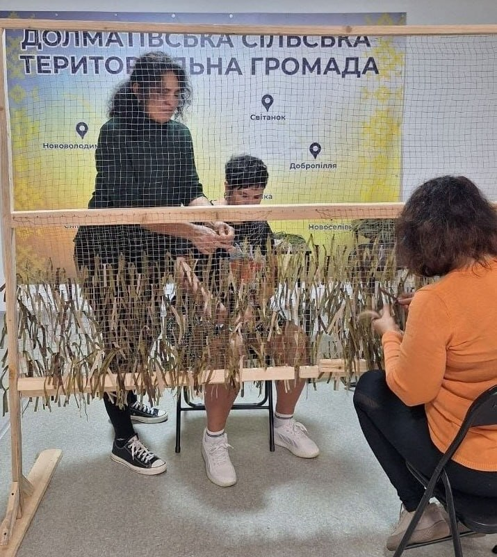 Волонтерки в координаційному центрі «Вільні разом» під час плетіння захисних маскхалатів для Захисників та Захисниць: на передньому плані на великому дерев'яному рамному станку натягнуто сітку, крізь яку три жінки зосереджено вплітають камуфляжні нитки та смужки тканини захисних природних відтінків. Ліворуч стоїть темноволоса жінка у чорному гольфі, темних штанах та чорно-білих кедах, яка працює руками з верхньою частиною сітки; по центру за станком на стільці сидить інша волонтерка у темному одязі та білих кросівках, а праворуч на передньому плані спиною до камери сидить жінка в помаранчевій кофті й чорних лосинах, яка ножицями підрівнює елементи маскхалата. На задньому плані за ними на стіні встановлено великий офіційний біло-жовтий банер із картою-схемою, відмітками населених пунктів (Нововолодимирівка, Світанок, Добропілля, Новоселівка тощо) та написом «ДОЛМАТІВСЬКА СІЛЬСЬКА ТЕРИТОРІАЛЬНА ГРОМАДА».