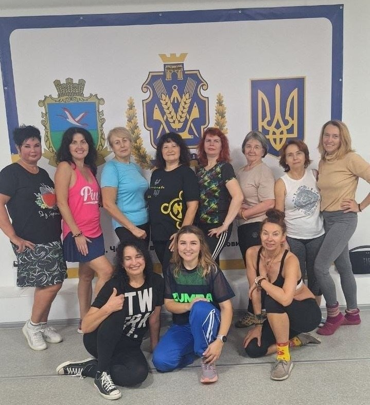 Спільне фото учасниць та тренерок після занять із зумби, йоги та стретчингу в Черкаському координаційному центрі «Вільні разом»: у світлій залі на тлі великого білого офіційного банера разом позує група жінок у різнокольоровому спортивному одязі (футболках, майках, легінсах та кросівках). На банері за ними зліва направо зображено символіку: герб Чаплинської громади (із білим птахом на синьо-зеленому тлі), герб Херсонської області по центру та державний тризуб України праворуч. Частина учасниць стоїть у верхньому ряду, поклавши руки на талію та посміхаючись у камеру, а попереду них на підлозі присіли ще три жінки, одна з яких показує жест «великий палець вгору», а інша одягнена у чорно-зелену футболку з написом «ZUMBA» та сині спортивні штани; усі присутні виглядають енергійно та задоволено після завершення тренування.