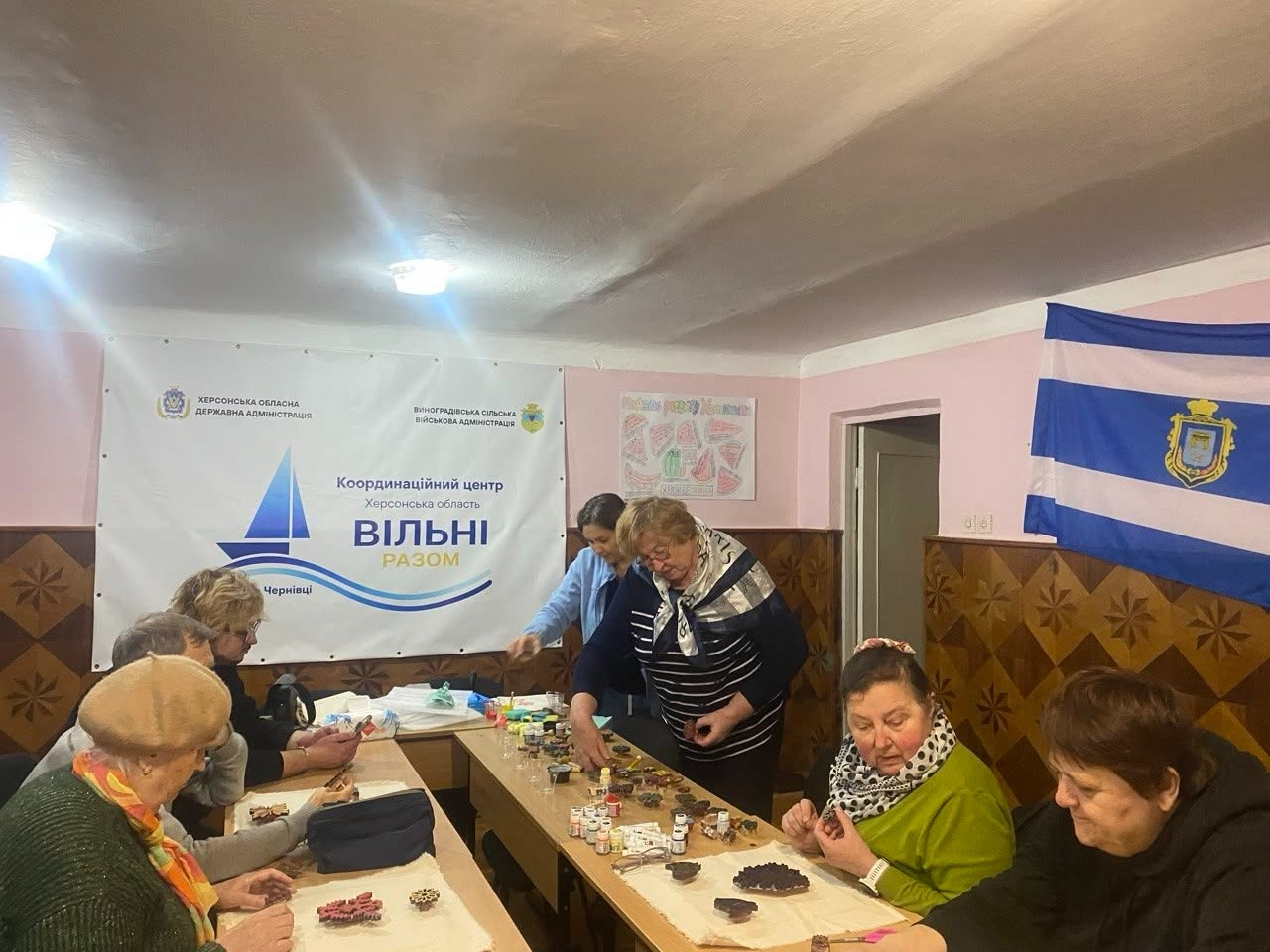 Майстер-клас із вибійки малюнків на тканині для переселенців із Херсонщини в Чернівецькому координаційному центрі «Вільні разом»: у кімнаті з декоративними дерев'яними панелями на стінах за довгим кутовим столом сидить група жінок старшого віку, які займаються творчістю. Дві майстрині-організаторки у центрі кадру допомагають учасницям, розкладаючи на столі фарби у тюбиках та дерев'яні штампи-вибійки різних форм. Жінки уважно обирають візерунки та наносять принти на біле тканинне полотно. На задньому плані ліворуч висить великий офіційний банер «Координаційний центр Херсонська область ВІЛЬНІ РАЗОМ у м. Чернівці» з логотипами Херсонської ОДА та Виноградівської сільської військової адміністрації. У крайньому правому кутку на стіні закріплено біло-синій прапор міста Херсон із гербом по центру.