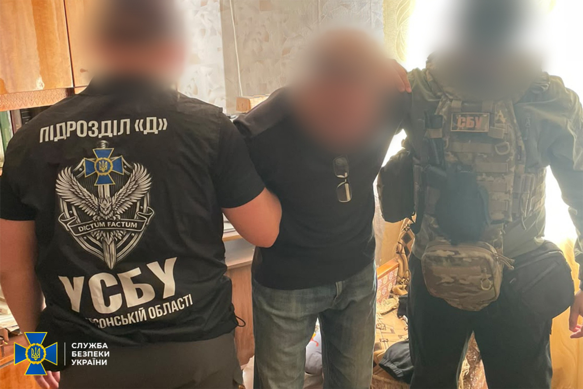 Затримання Службою безпеки України колаборанта в Херсоні: у кімнаті житлового будинку двоє бійців СБУ утримують під руки затриманого чоловіка, чиє обличчя приховане цифровим розмиттям; ліворуч стоїть оперативник у чорній спецформі, на спині якої нанесено великі білі написи «ПІДРОЗДІЛ «Д» та «УСБУ В ХЕРСОНСЬКІЙ ОБЛАСТІ», а також шеврон із мечем та девізом «DICTUM FACTUM»; праворуч тримає за плече інший боєць у камуфляжній формі та розвантажувальному жилеті з емблемою «СБУ», чиє обличчя так само закрите маскою; затриманий посередині чоловік одягнений у чорну футболку із сонцезахисними окулярами на комірі та сині джинси; у лівому нижньому кутку кадру розміщено офіційний логотип Служби безпеки України.
