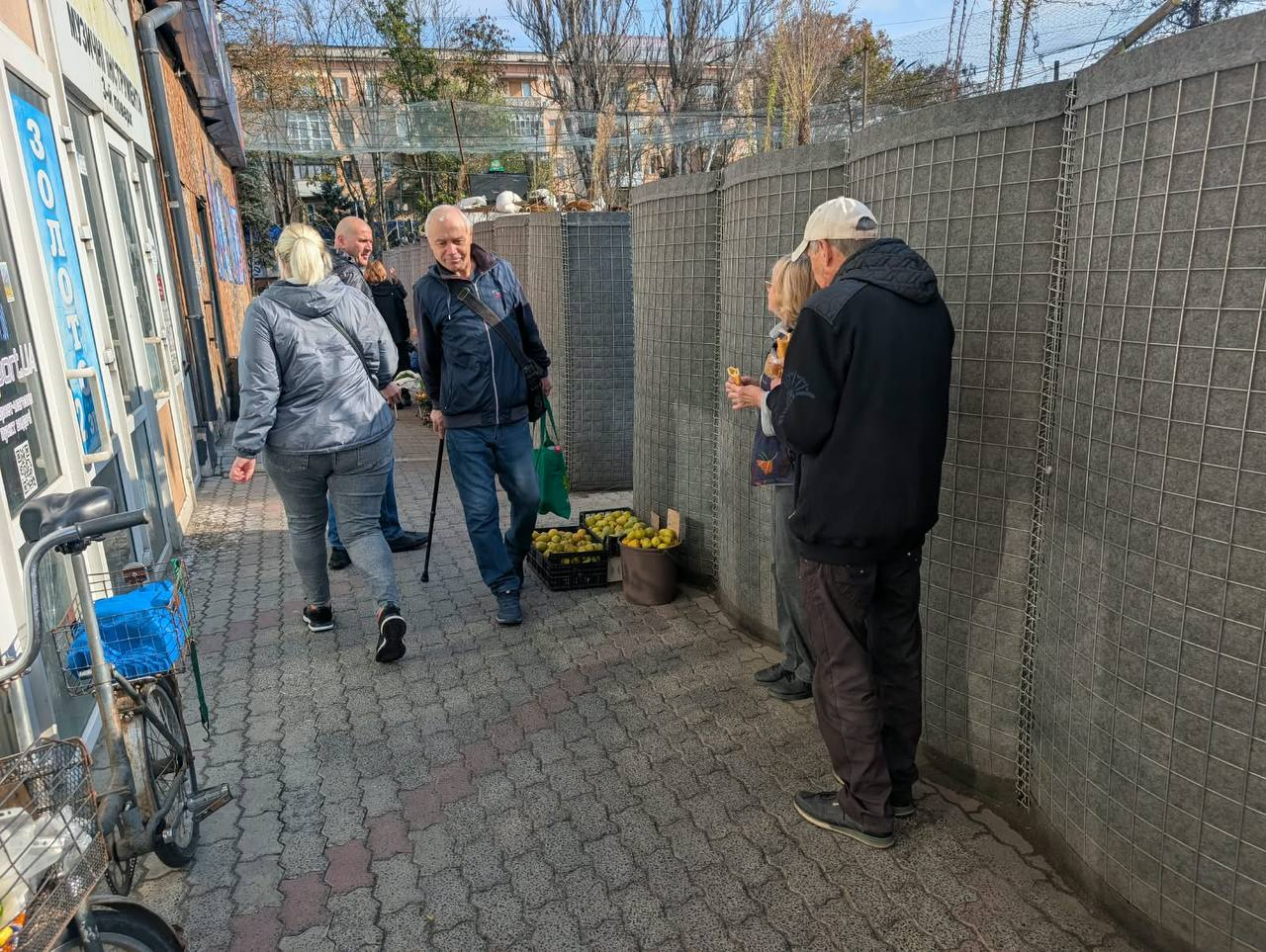 Місцеві жителі на одній із вулиць Херсона під час дії режиму припинення вогню та моніторингу дотримання тиші: на викладеній бруківкою пішохідній зоні біля торговельних павільйонів спілкуються та прогулюються кілька людей похилого віку й перехожих. По центру кадру літній чоловік у синій куртці, джинсах та з чорною сумкою через плече посміхається, спираючись на ортопедичний ціпок-палицю і тримаючи в лівій руці зелений екопакет. Ліворуч від нього спиною до камери йде жінка у сірій куртці та сірих джинсах, а на задньому плані видно ще одного чоловіка. Праворуч, біля високого захисного паркана-габіону зі зварної сітки та каменю, стоїть пара — чоловік у чорній куртці та світлій бейсболці й жінка, які розмовляють та їдять фрукти. Поруч із ними на землі стоять пластиковий ящик та відро, вщент наповнені стиглою жовтою айвою або яблуками, а в крайньому лівому кутку помітно припаркований старий велосипед із синім кошиком.