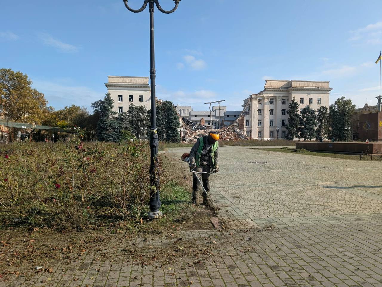 Комунальні роботи на площі Свободи у Херсоні під час дії режиму тиші та припинення вогню: на передньому плані по центру чоловік-комунальник у захисному шоломі, навушниках та зеленому сигнальному жилеті косить траву ручним бензиновим тримером біля газону та високого чорного металевого ліхтаря; площа викладена сірою бруківкою, на якій лежить скошена трава та сухе осіннє листя. На задньому плані через усю площу видно монументальну будівлю Херсонської ОДА, центральна частина якої повністю зруйнована внаслідок бойових дій, утворивши величезний пролом із руїнами з бетонних плит та цегли; праворуч на площі піднято на флагштоку державний прапор України, а небо над містом безхмарне та синє.