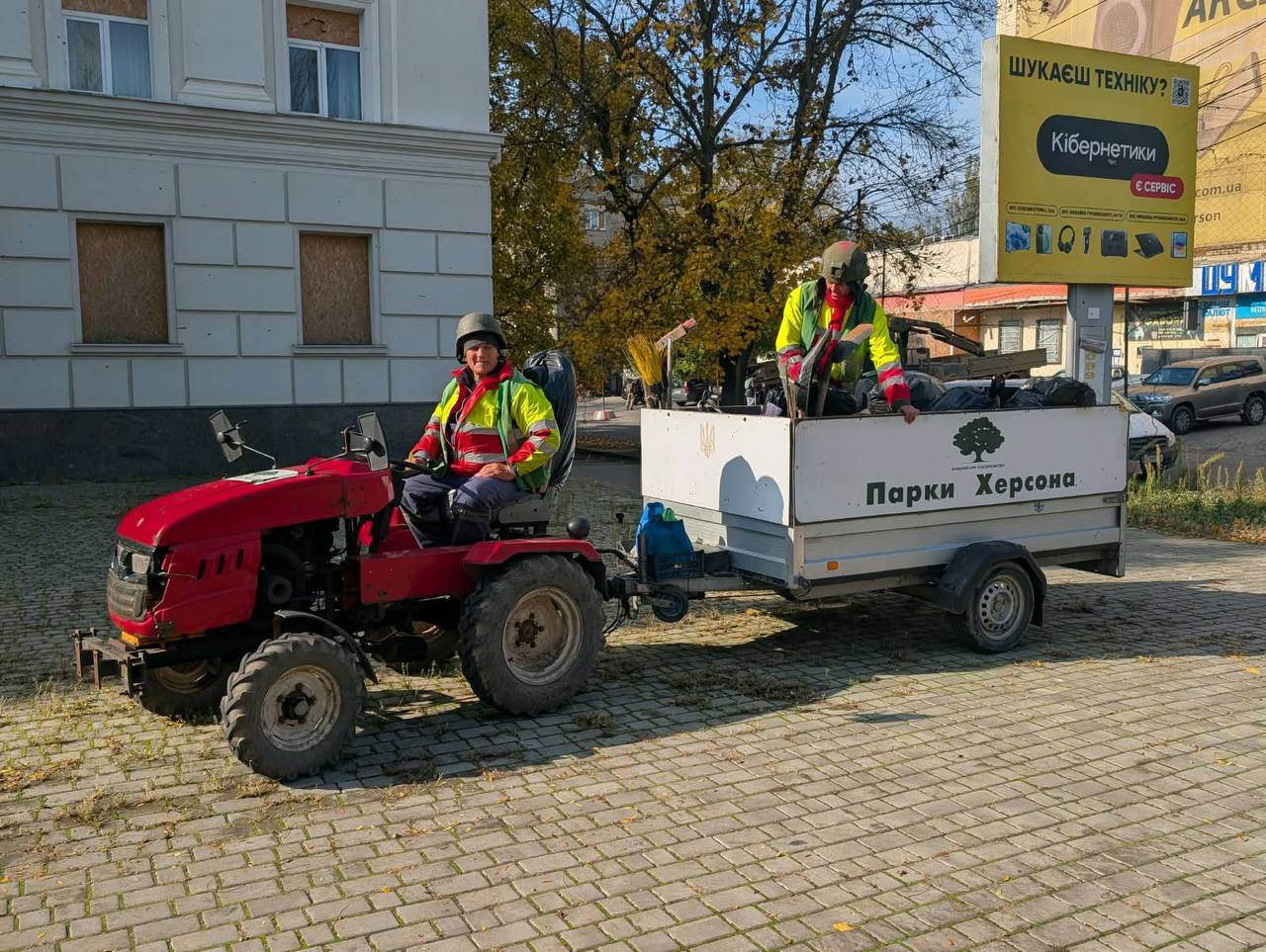 Робота комунальних служб КП «Парки Херсона» на вулицях міста під час дії режиму тиші та припинення вогню: по центру на викладеній бруківкою дорозі зафіксовано невеликий червоний мінітрактор з автомобільним причепом, за кермом якого у захисному військовому шоломі та сигнальному яскраво-зеленому жилеті зі світловідбивними смугами сидить чоловік-водій і посміхається в камеру. У білому металевому причепі, на борту якого нанесено фірмовий логотип підприємства з деревом, золотий герб України та напис «Парки Херсона», стоїть інший комунальник у касці й збирає сміття та сухе листя у великі чорні мішки за допомогою мітел та лопат. На задньому плані ліворуч височіє старовинна світла триповерхова будівля, вікна першого поверху якої забиті дерев'яними ОСБ-плитами для захисту від обстрілів; праворуч над причепом встановлено великий жовтий рекламний білборд магазину «Кібернетики» із закликом «Шукаєш техніку?», а в глибині вулиці за деревами помітно припаркований позашляховик та інші міські споруди.