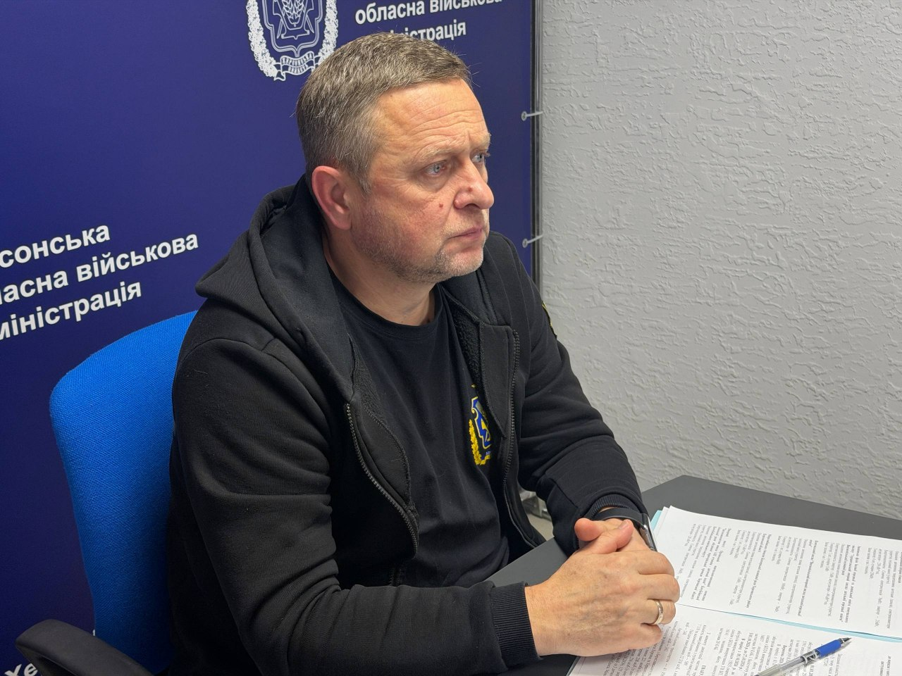 Перший заступник начальника Херсонської ОВА Дмитро Бутрій під час онлайн-наради з питань евакуації та розселення населення: у кабінеті за сірим робочим столом сидить у профіль праворуч посадова особа — чоловік середнього віку з короткою стрижкою, сивиною на скронях та легкою щетиною; він одягнений у чорну трикотажну кофту на блискавці з капюшоном, під якою видно чорну футболку з маленьким жовто-синім гербом України на грудях. Чоловік сидить у синьому офісному кріслі, зосереджено дивиться перед собою в бік і тримає руки на столі в замку, де перед ним лежать офіційні друковані документи, бланки та синя кулькова ручка. На задньому плані ліворуч встановлено великий темно-синій інформаційний банер із білими офіційними написами «Херсонська обласна військова адміністрація» та зображенням герба області у верхній частині, а права стіна приміщення має світло-сіру фактурну штукатурку.