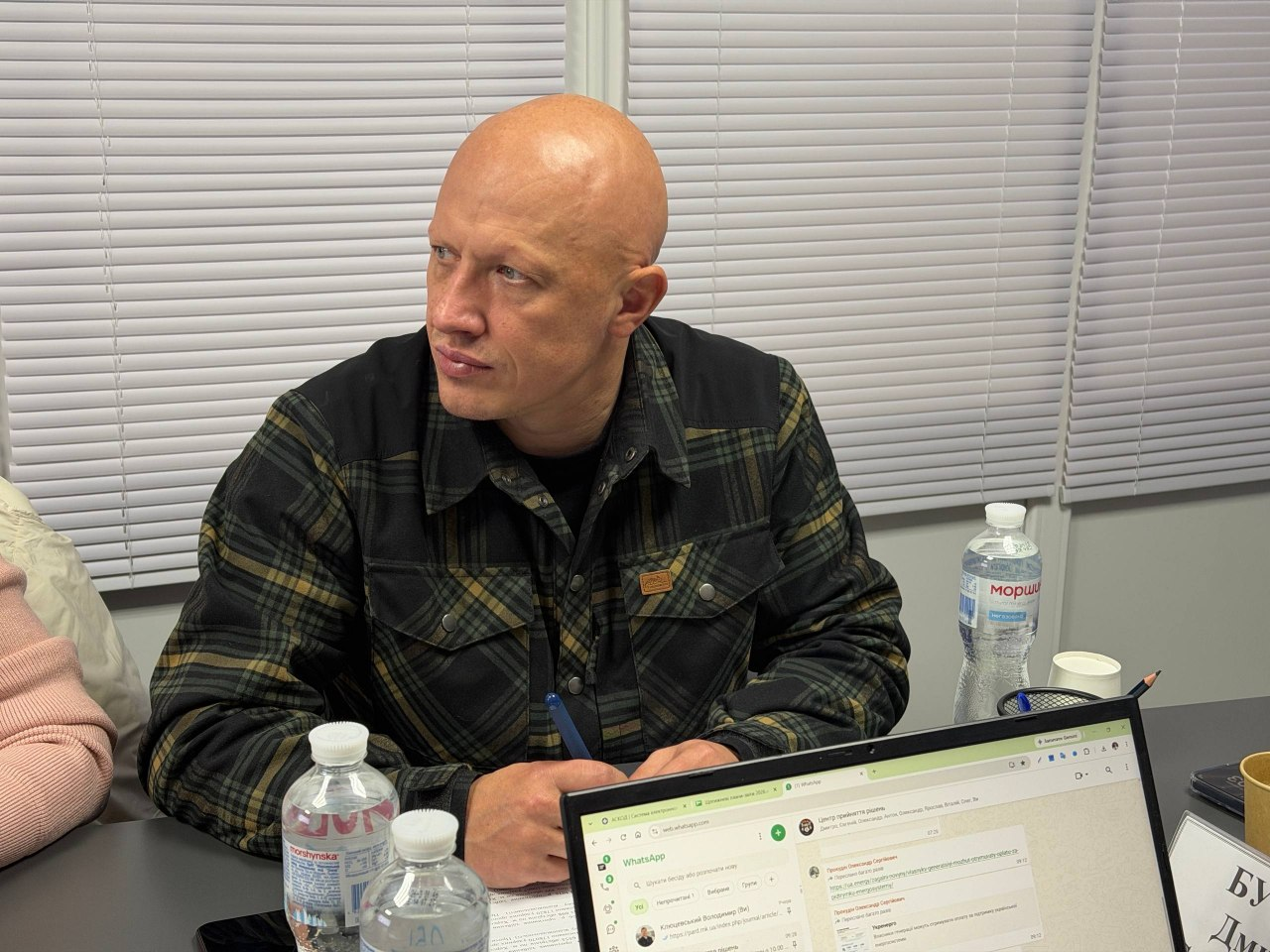 Заступник начальника Херсонської ОВА Євгеній Ігнатенко під час онлайн-наради з питань евакуації та розселення населення: у кабінеті за робочим столом сидить чоловік міцної статури, повернутий напівпрофілем ліворуч, який уважно слухає хід обговорення; він одягнений у щільну теплу сорочку в темно-зелену та чорну клітинку з двома нагрудними кишенями на ґудзиках, під якою видно чорну футболку. Перед посадовцем на столі стоять дві пластикові пляшки мінеральної води «Моршинська» з білими кришками, а безпосередньо на передньому плані розташований відкритий ноутбук, на екрані якого відображено робоче вікно месенджера WhatsApp із відкритими чатами та повідомленнями; праворуч на столі помітно органайзер для канцелярії з олівцями, паперовий стаканчик та край настільної таблички. На задньому плані вікно приміщення повністю завішене світлими горизонтальними офісними жалюзі.
