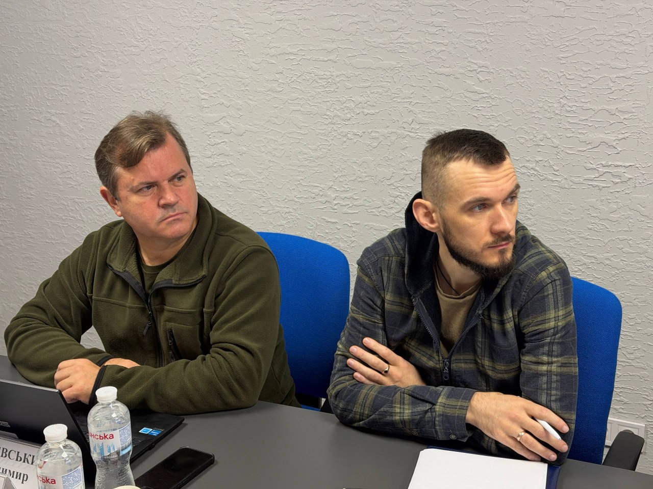 Заступник голови Херсонської ОДА Володимир Клюцевський та заступник директора Департаменту здоров’я Херсонської ОДА Михайло Балацький під час онлайн-наради з питань евакуації та розселення населення: за довгим робочим столом на синіх офісних кріслах сидять двоє чоловіків, повернутих напівпрофілем праворуч, які уважно стежать за ходом обговорення на екрані. Ліворуч сидить посадова особа — заступник голови Херсонської ОДА Володимир Клюцевський у військовій оливковій флісовій кофті, перед яким на столі лежить закритий чорний ноутбук, мобільний телефон та стоїть пластикова пляшка мінеральної води «Миргородська». Праворуч від нього сидить заступник директора Департаменту здоров’я Херсонської ОДА Михайло Балацький із короткою стрижкою та акуратною темною бородою, одягнений у щільну сорочку в синьо-зелену клітинку з капюшоном, який схрестив руки перед собою, тримаючи в пальцях білу ручку над розгорнутими паперовими документами. На задньому плані кімнати стіна має однотонну світло-сіру фактурну штукатурку.