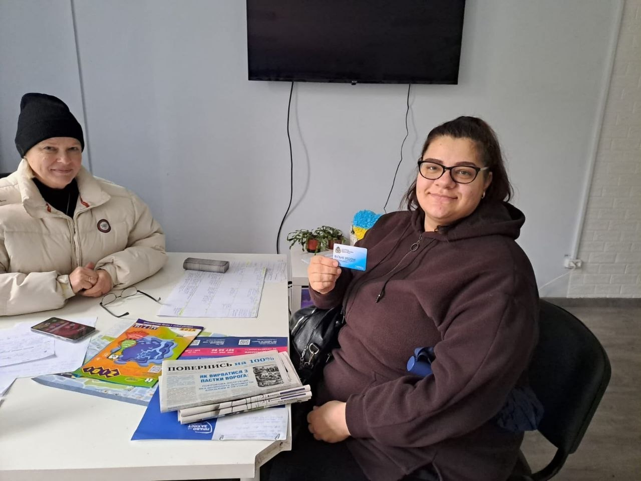 Видача карток у межах проєкту «Картка Херсонщини» для переселенців у Львівському координаційному центру «Вільні разом»: у світлому кабінеті за білим кутовим столом сидять дві жінки; ліворуч сидить представниця хабу у світлій куртці-пуховику та чорній шапці, яка склала руки перед собою; праворуч на стільці сидить дівчина-переселенка в окулярах та темно-коричневому худі з капюшоном, яка посміхається та тримає у правій руці отриману синьо-блакитну пластикову «Картку Херсонщини». На столі перед ними розкладено робочі папери, бланки, лежать сонцезахисні окуляри, смартфон та стос свіжих газет із заголовком «ПОВЕРНИСЬ на 100%. ЯК ВИРВАТИСЯ З ПАСТКИ ВОРОГА». На задньому плані на світло-сірій стіні висить великий вимкнений плазмовий телевізор, під яким на тумбі помітно невеликі кімнатні рослини та патріотичний синьо-жовтий декор.