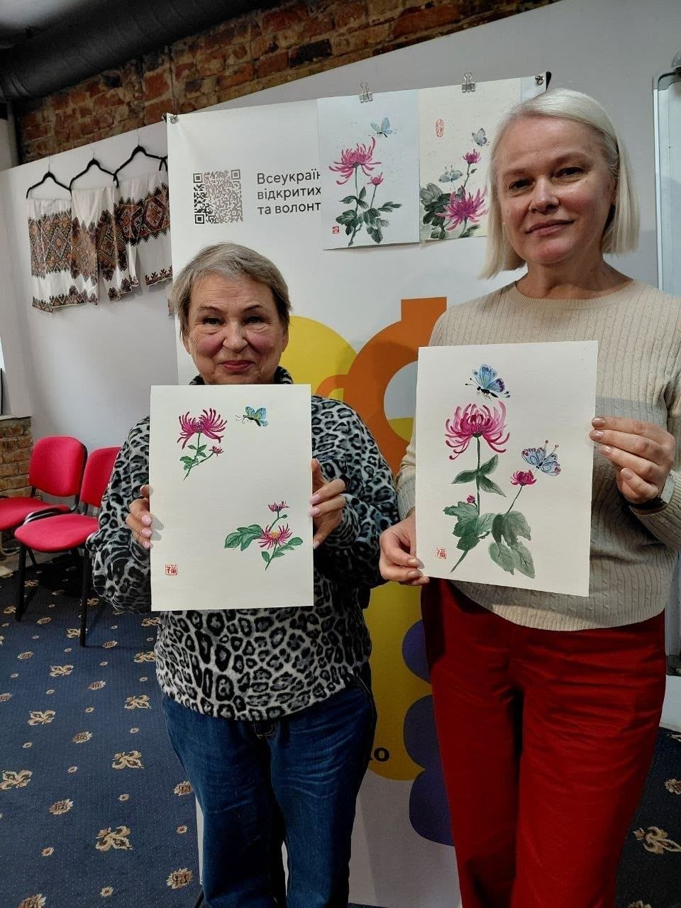 Дві учасниці майстер-класу з китайського живопису від координаційного центру «Вільні разом» посміхаються та тримають у руках свої готові малюнки із зображенням рожевих хризантем і метеликів. Жінка ліворуч має коротку зачіску з русяво-сивим волоссям, вона одягнена в кофту з леопардовим принтом і сині джинси, а жінка праворуч — із зачіскою каре та світлим волоссям у світлому бежевому светрі та яскраво-червоних штанах. На вертикальних аркушах білого паперу, які вони демонструють, детально промальовані квіти, зелене листя, блакитні та рожеві метелики, а також поставлені маленькі червоні відбитки авторських печаток. На задньому плані видно стіну з плакатом «Всеукраїнська мережа відкритих та волонт...» і QR-кодом, зразки інших художніх робіт, вишиванки на вішаках, червоні офісні стільці та темний килим із візерунком на підлозі.