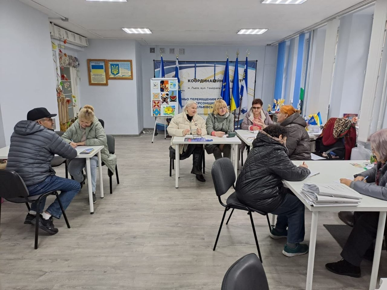 У світлому кабінеті координаційного центру «Вільні разом» у Львові переселенці з Херсонщини отримують індивідуальні консультації за столами. У приміщенні облаштовано кілька робочих зон: ліворуч чоловік та жінка в куртках спілкуються з фахівцем, у центрі відвідувачі сидять навпроти фахівців, а праворуч жінка-консультант розбирає папери та надає роз'яснення відвідувачкам. На задньому плані кімнати встановлено великий інформаційний банер із синьо-жовтою символікою та текстом «Координаційний центр... допомоги переміщеним... з Херсонщини...», перед яким стоять три великі прапори на підставках, серед яких видніється Державний Прапор України та крайній праворуч — прапор Херсонської області. Ліворуч від банера стоїть білий інформаційний стенд-банер із різнокольоровими дитячими малюнками, за яким стоять ще два прапори Херсонської області на підставках. На стіні ліворуч розміщені в рамках Державний Герб України та Державний Прапор України, а з правого боку кімнати видно великі вікна з вертикальними жалюзі.