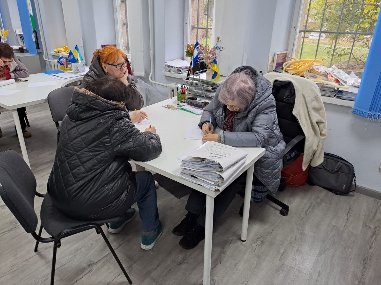 У світлому кабінеті координаційного центру «Вільні разом» у Львові відвідувачі з Херсонщини отримують індивідуальні консультації за робочим столом. На передньому плані ліворуч сидять дві відвідувачки в темних куртках, одна з яких має яскраво-руде волосся та окуляри, і уважно спостерігають за процесом. Навпроти них за білим столом сидить жінка-консультант у сірій куртці та окулярах, яка робить записи на аркуші паперу поруч із великим стосом документів. На задньому плані кімнати видно інший робочий стіл, великі вікна з вертикальними жалюзі, папки з паперами, а також патріотичну символіку — маленькі прапорці України.