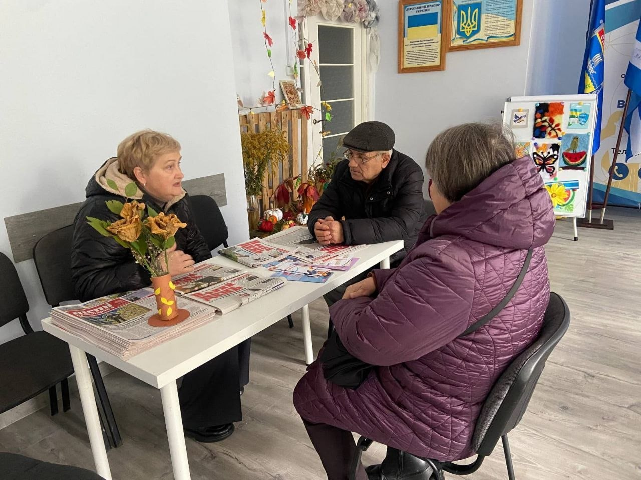 У приміщенні координаційного центру «Вільні разом» у Львові відвідувачі з Херсонщини зібралися за столом для отримання свіжих номерів газети «Новий день». Ліворуч за довгим білим столом сидить жінка в чорній куртці з капюшоном, яка спілкується з гостями, а поруч із нею стоїть вазочка з паперовими квітами та розкладені великі стоси газет. Навпроти неї та на торці столу сидять літній чоловік у чорній куртці й кепці та жінка в об'ємній фіолетовій куртці спиною до камери. На задньому плані кабінету на стіні розміщені в рамках Державний Герб України та Державний Прапор України, поруч стоїть мольберт із різнокольоровими малюнками, а праворуч видніються два великі прапори на підставках, де крайнім праворуч є прапор Херсонської області.