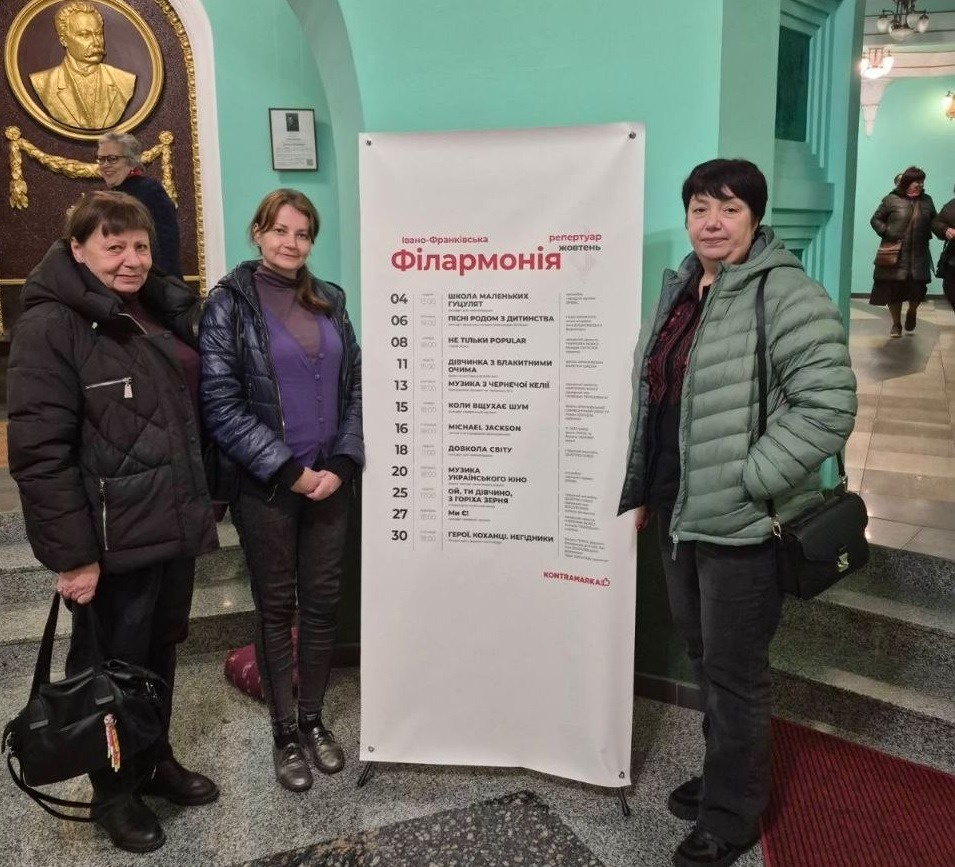 Три жінки з числа переселенців із Херсонщини позують у фоє Івано-Франківської обласної філармонії імені Іри Маланюк біля великого білого банера-афіші. Жінка ліворуч одягнена в подовжену чорну зимову куртку, темні штани, тримає в руці чорну сумку і посміхається. Поруч із нею стоїть жінка у фіолетовому кардигані, сірому гольфі, темних облягаючих штанах і високих черевиках, склавши руки перед собою. Праворуч від банера стоїть третя відвідувачка в об'ємній світло-зеленій куртці, чорних штанах та з чорною сумкою через плече. Великий вертикальний банер містить напис «Івано-Франківська філармонія. Репертуар жовтень» та розклад концертних програм із датами, серед яких вказано і концерт «Ми є». На задньому плані видно світло-зелені стіни, колони, сходи, круглий золотистий барельєф із портретом на стіні ліворуч та інших відвідувачів у фоє закладу.